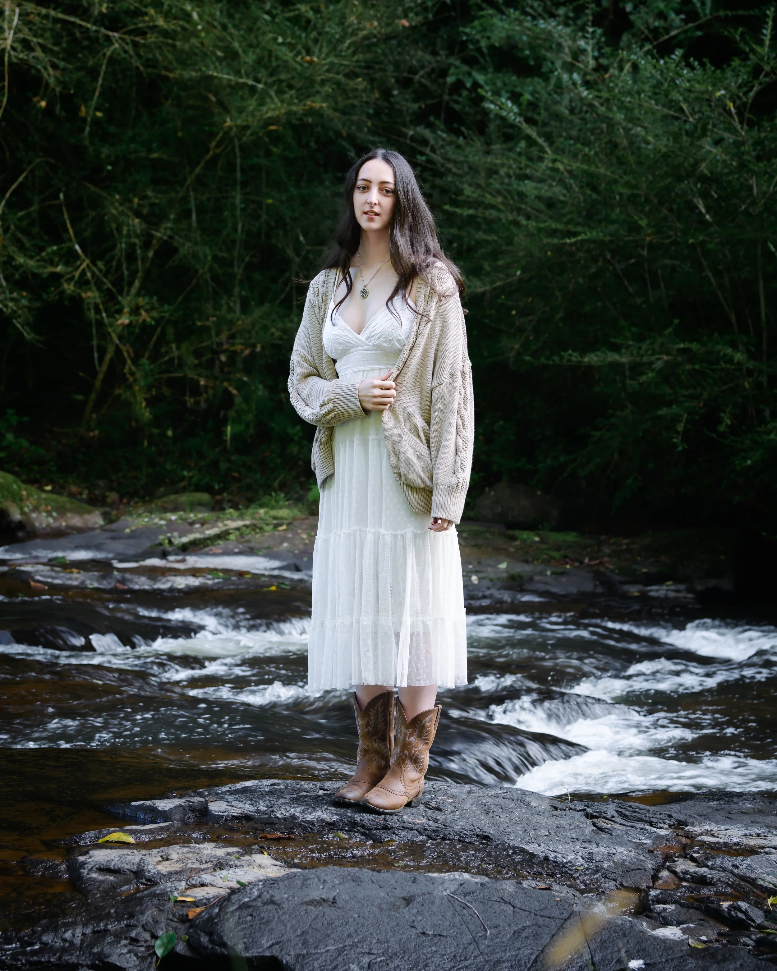Shanleigh Rose in a white dress, tan cowboy boots, and a beige cardigan standing on a rock in a shallow stream, with lush green foliage in the background.