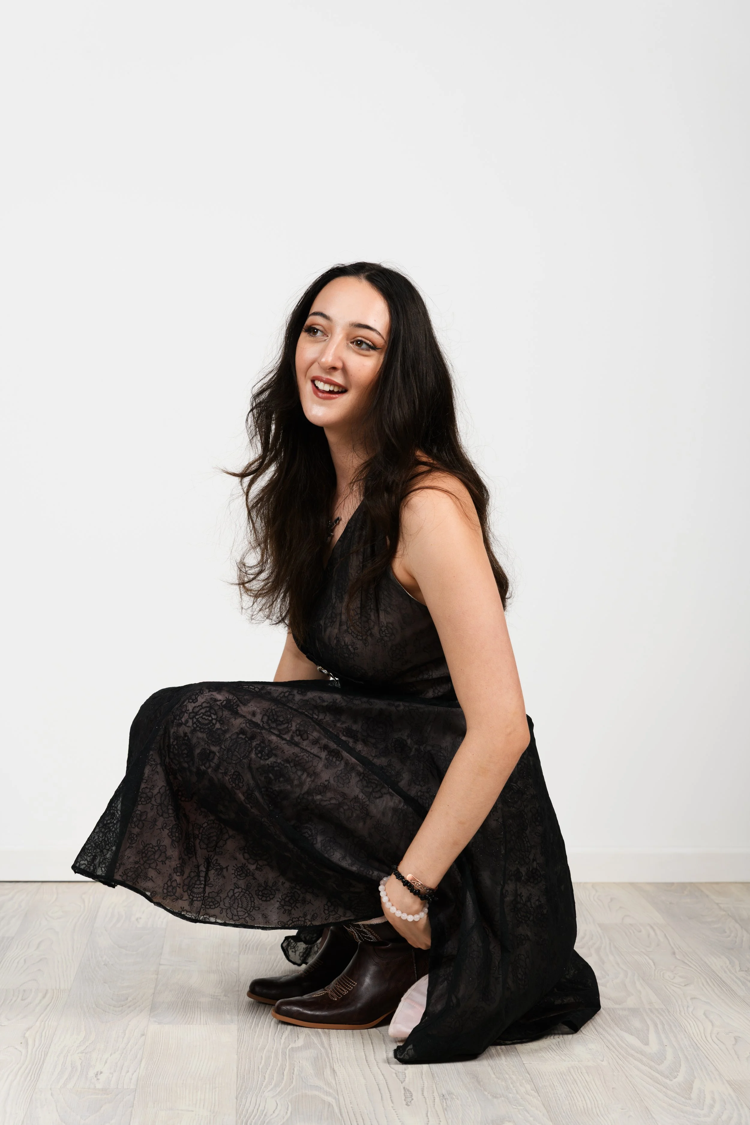 Shanleigh Rose with long dark hair, wearing a black lace dress, crouching down with a smile, adjusting her brown boots.