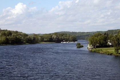 A river flowing through a green landscape with trees and grassy areas on both sides under a partly cloudy sky.