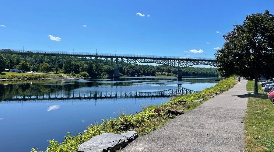 A scenic riverside view with a bridge crossing over the water, a sidewalk along the bank with parked cars on one side, and trees providing shade, under a partly cloudy blue sky.