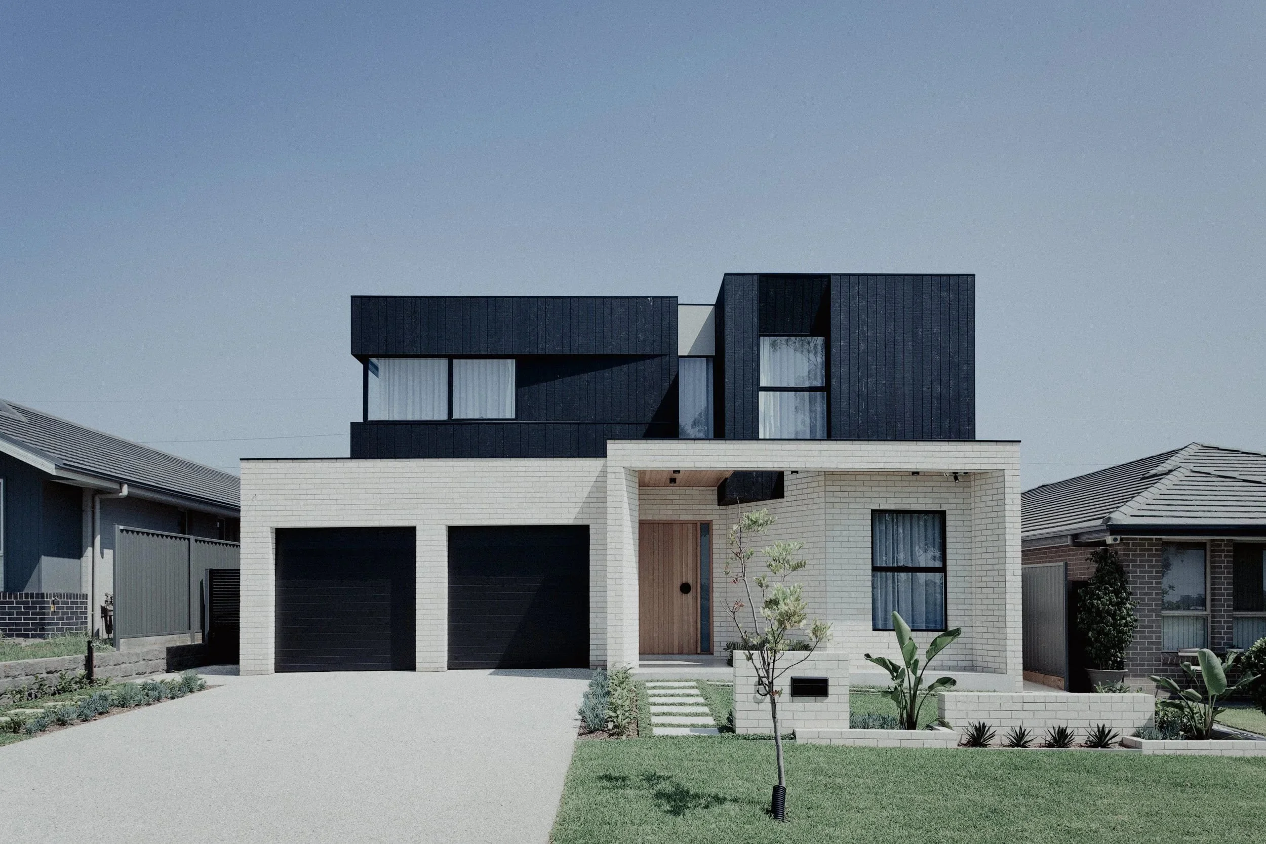 Modern two-story house with white brick and black upper story, black garage doors, and a small front yard with landscaping and a young tree.