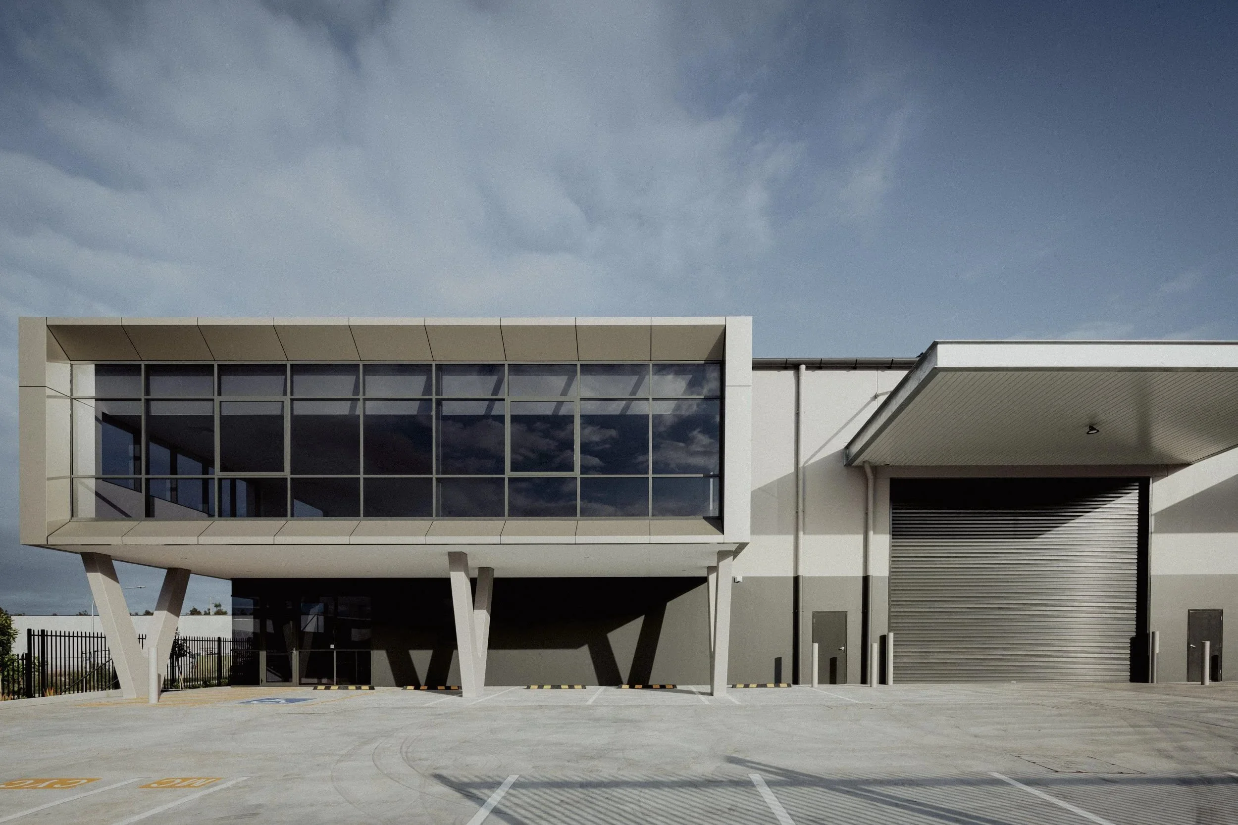 Modern industrial building with a glass-fronted section on stilts and a large roller door, set in a parking lot under a partly cloudy sky.