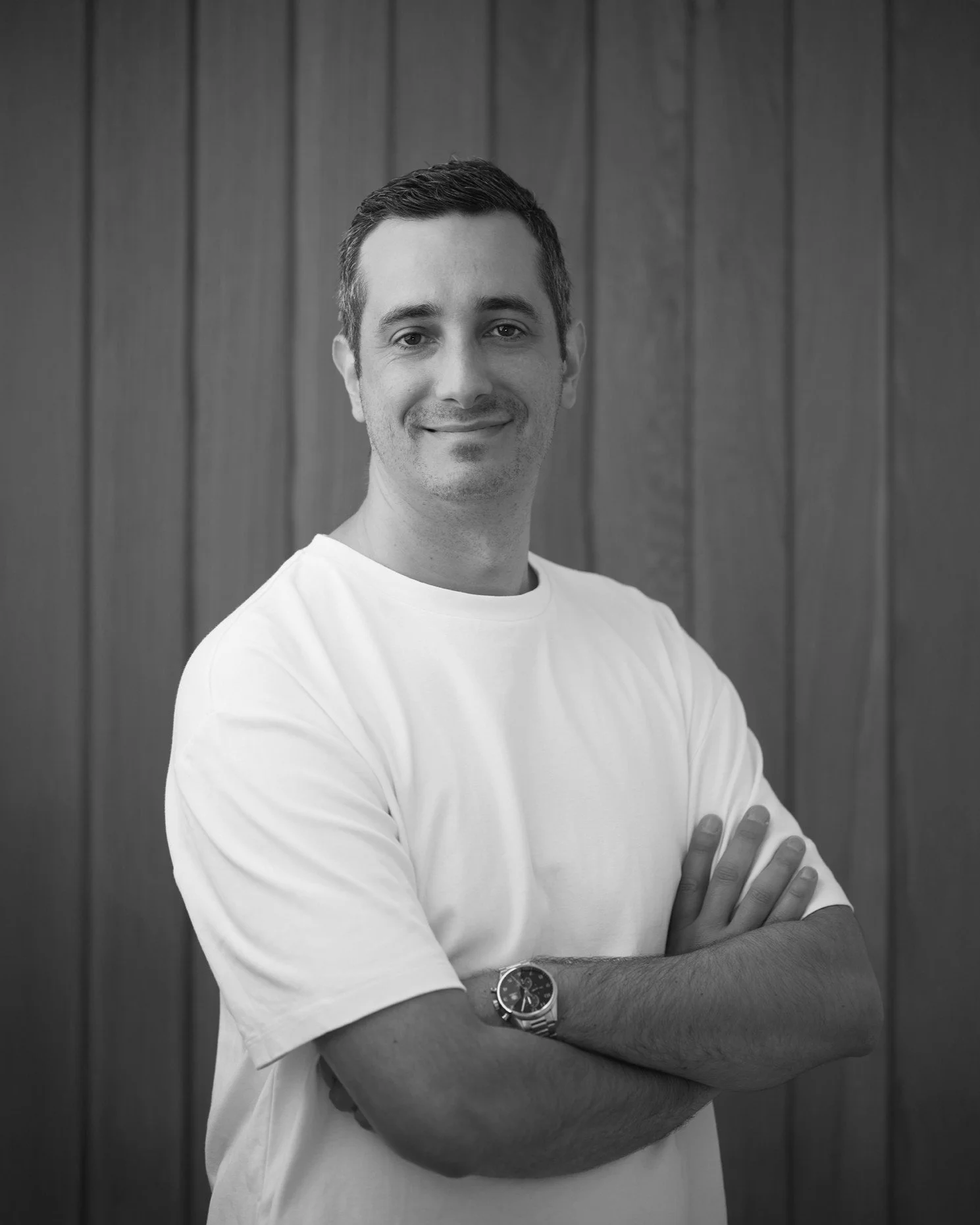 Black and white portrait of a man with short hair, smiling, wearing a white T-shirt and a watch, standing with arms crossed in front of a wooden wall.