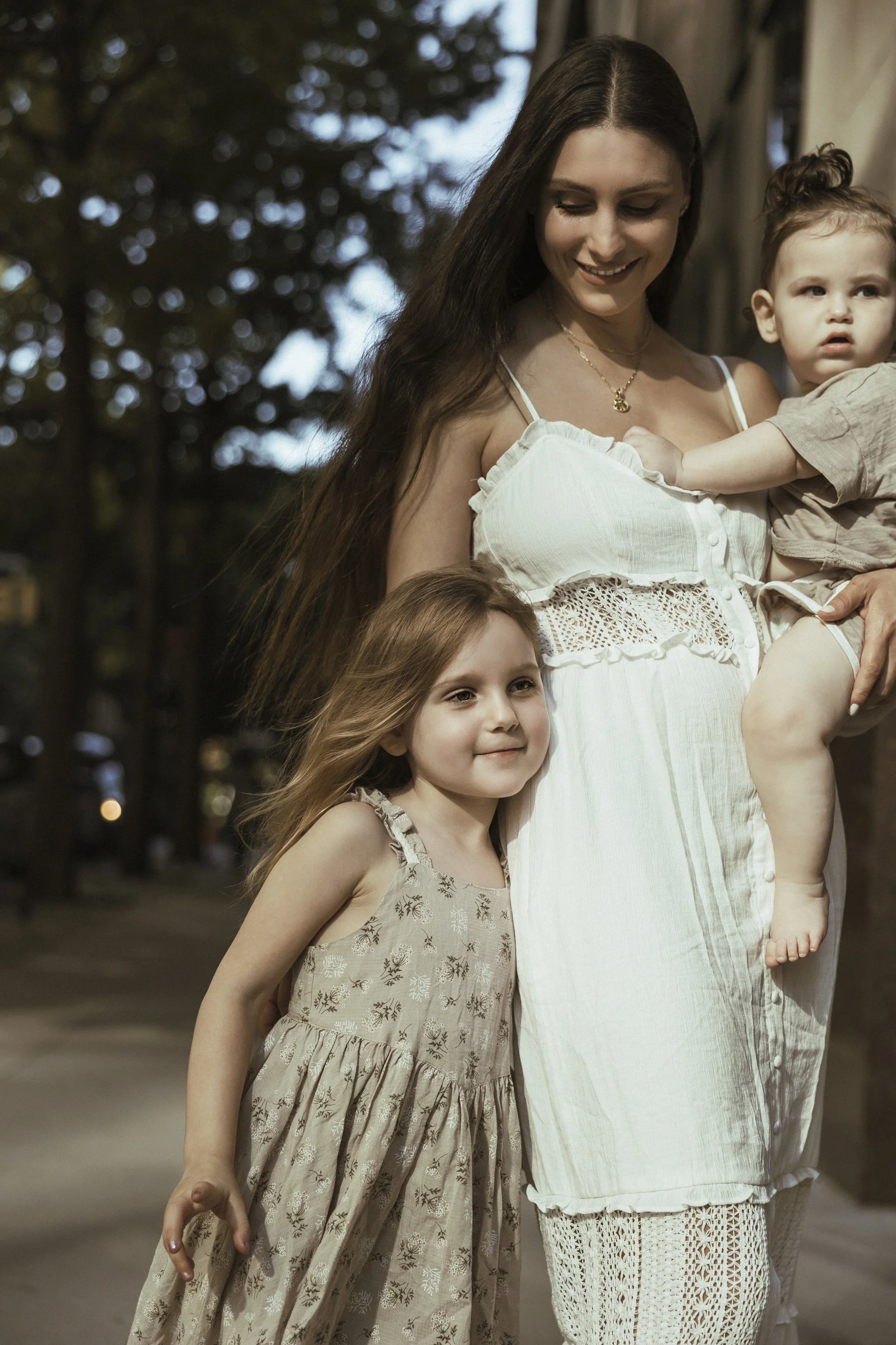 A woman in a white dress holding a young girl and a young boy, standing outdoors during the day. The woman has long dark hair, and the children have light brown hair. The girl is smiling, and the boy is looking to the side.