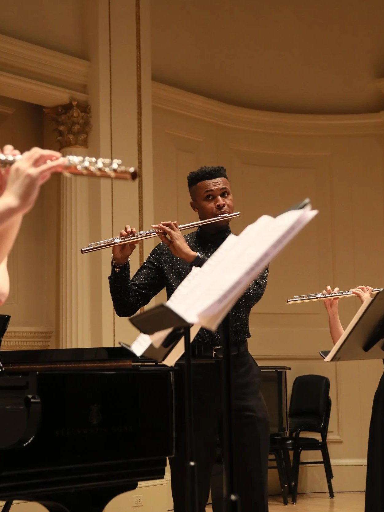 A young man with a short fade haircut playing the flute during a concert, surrounded by other musicians, in a formal concert hall with ornate decor.