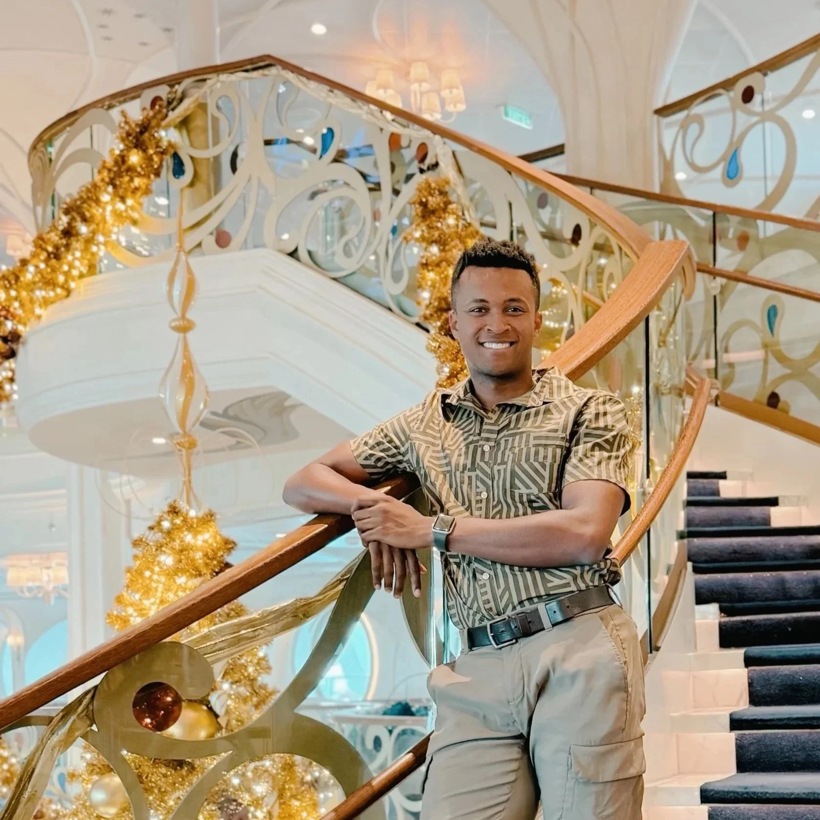 A young man standing on a stairway in a decorated, luxurious interior with gold Christmas trees and ornate railing, smiling at the camera.