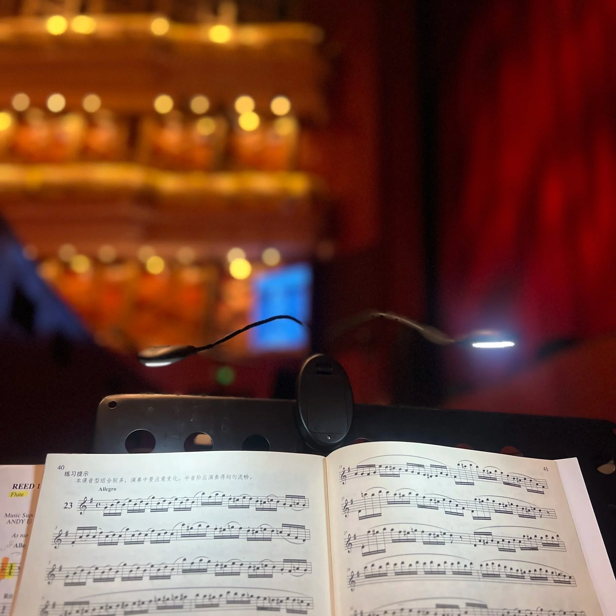 Close-up of a sheet of music for flute, with music notes and Chinese characters, resting on a music stand in a dimly lit room with blurred golden and red background lights.
