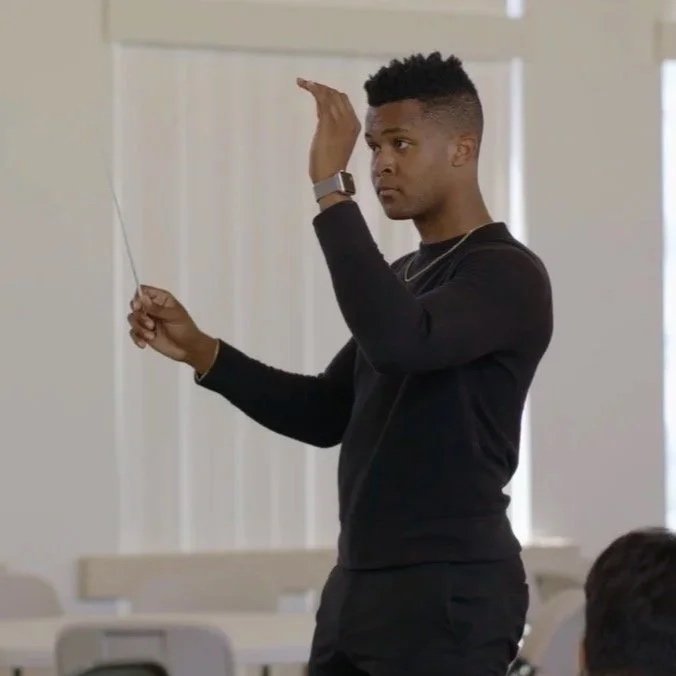 A young man in black clothing holding a pointer, raising his hand, and looking attentively during a presentation or lecture in a classroom or conference room.