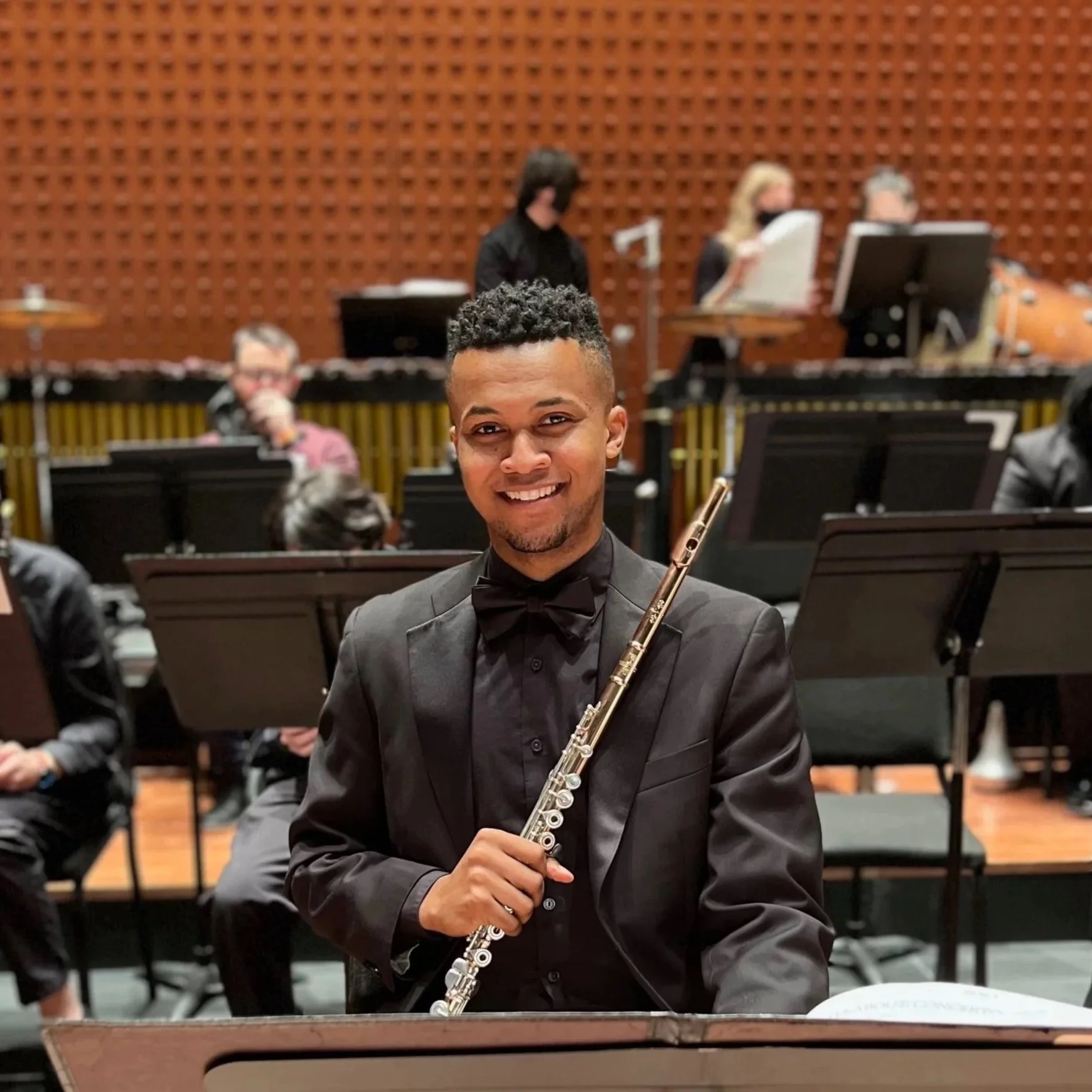 A young man in a tuxedo holding a flute, seated in front of an orchestra with musicians and music stands in a concert hall.
