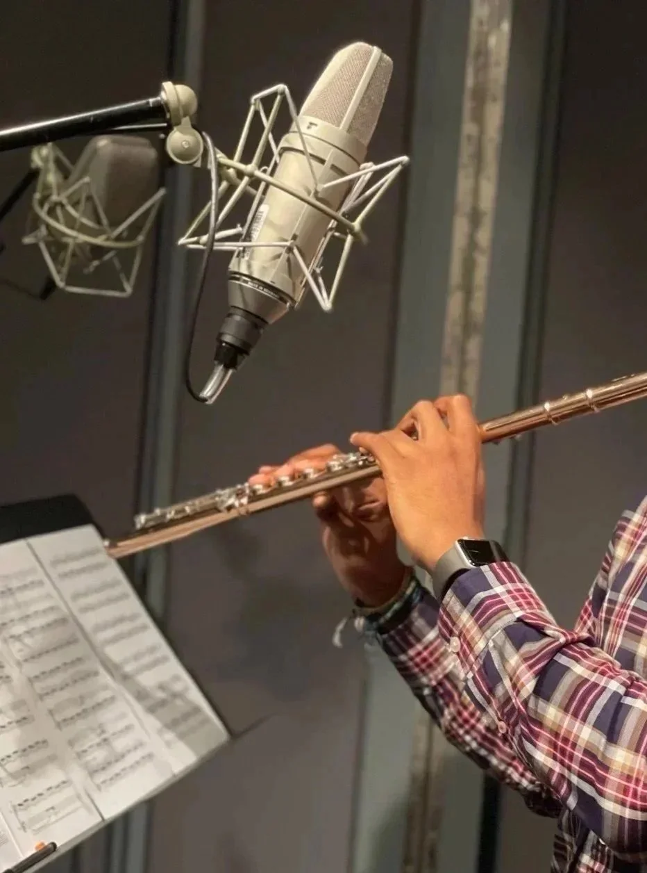 A person playing a silver flute in a recording studio with a microphone positioned above the instrument and sheet music in front.