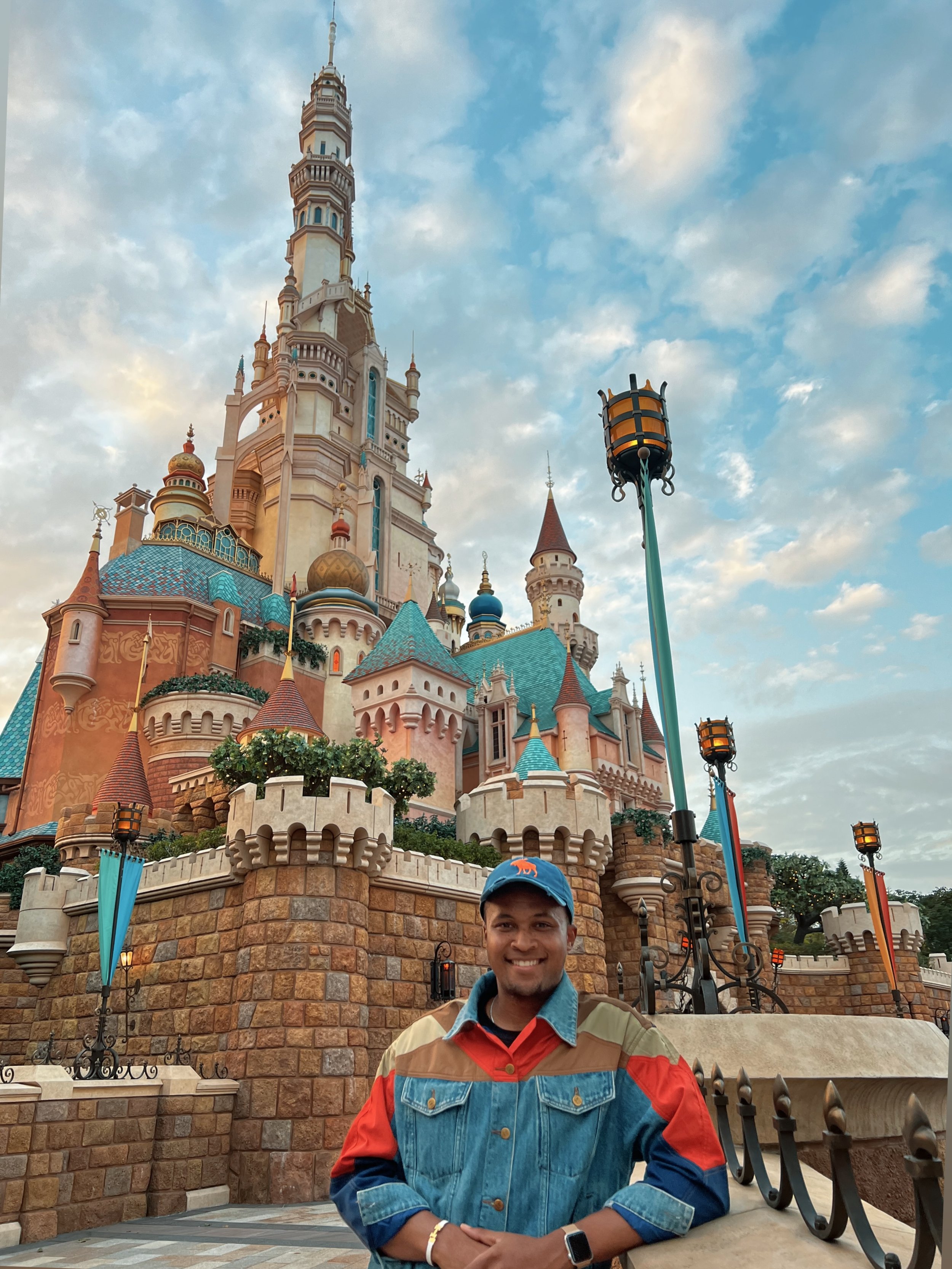 Dennis Rendleman smiling in a colorful jacket and blue cap standing in front of the Enchanted Storybook Castle at Disneyland Hong Kong during sunset.