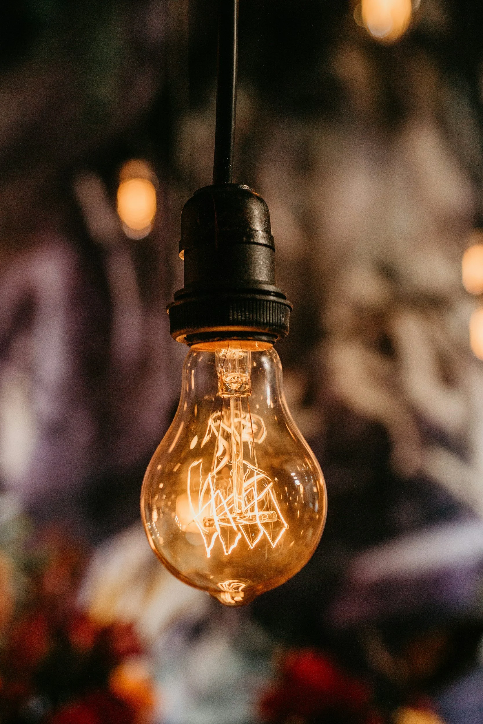 Close-up of an illuminated Edison-style light bulb hanging against a dark background with blurred lights and objects.