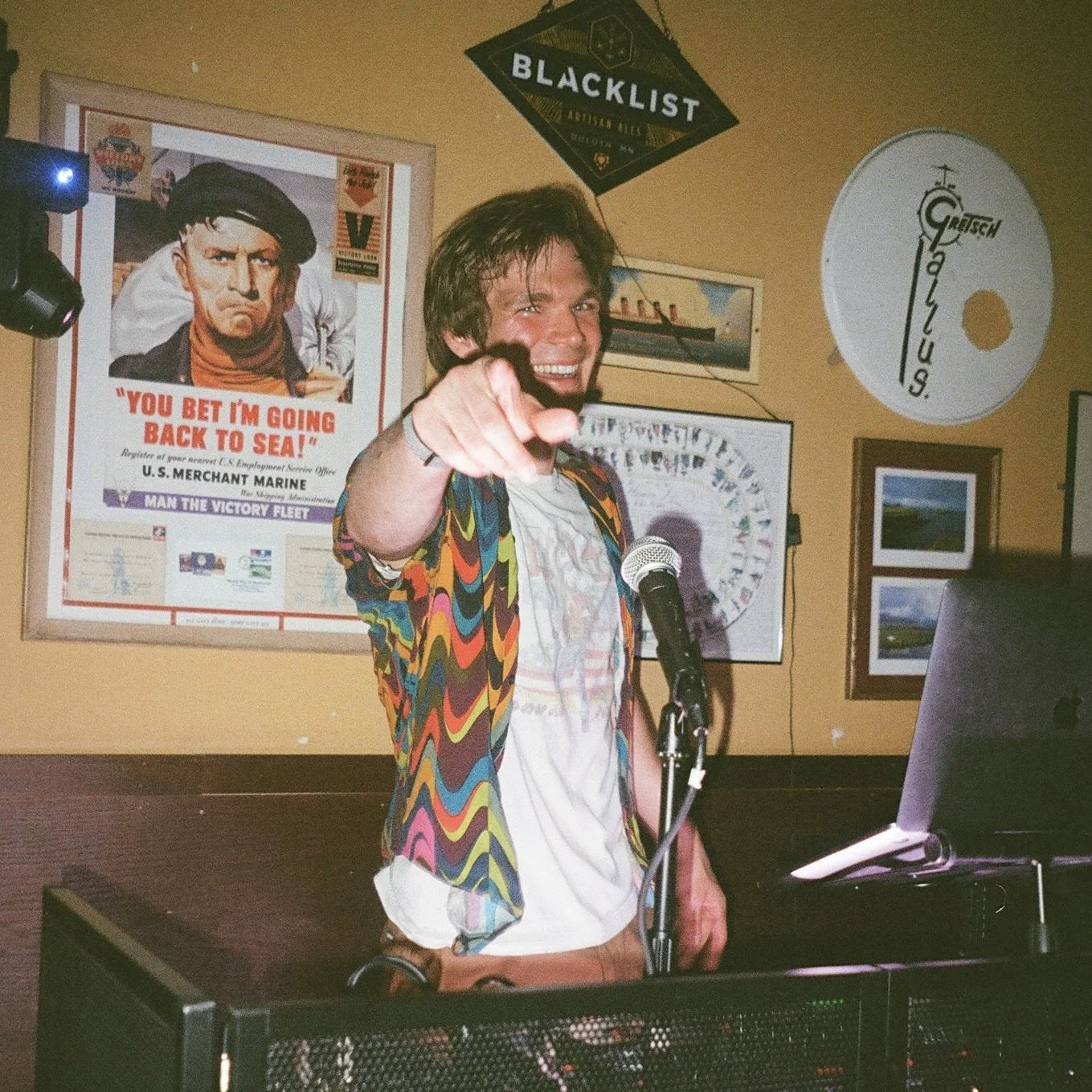 A man with wavy brown hair wearing a colorful, zigzag patterned jacket, white shirt, and brown pants, smiling and pointing towards the camera. He is standing behind a microphone with a keyboard and electronic equipment in front of him inside a room decorated with framed pictures, posters, and a 45 RPM vinyl record wall decoration.