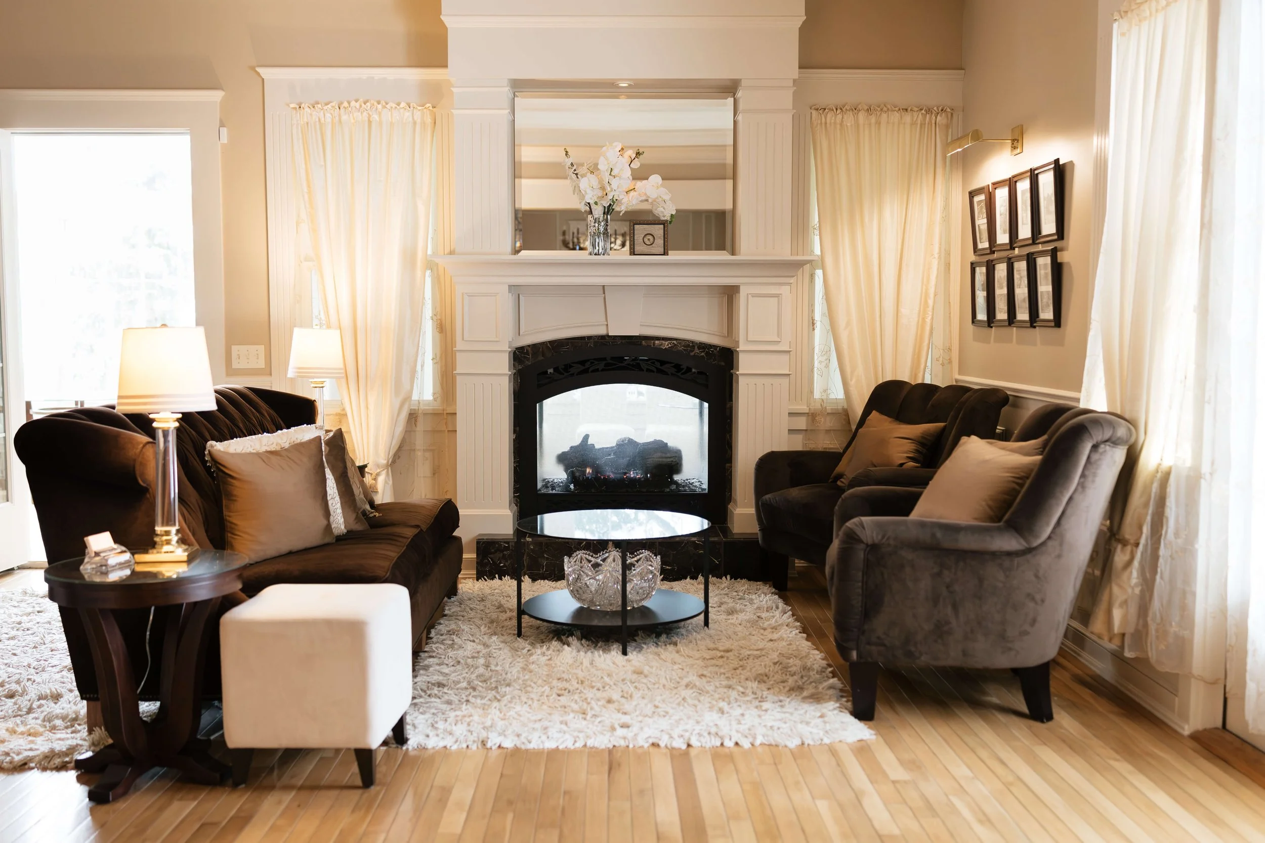 Living room with a white fireplace, a mirror above the fireplace, a vase of white flowers, brown and gray sofas, table lamps, a small round glass coffee table, framed pictures on the wall, and curtains on large windows.