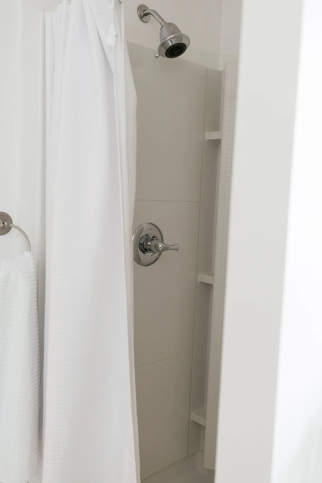 A shower stall with a silver showerhead, a silver faucet handle, a white shower curtain, and some built-in shelves.
