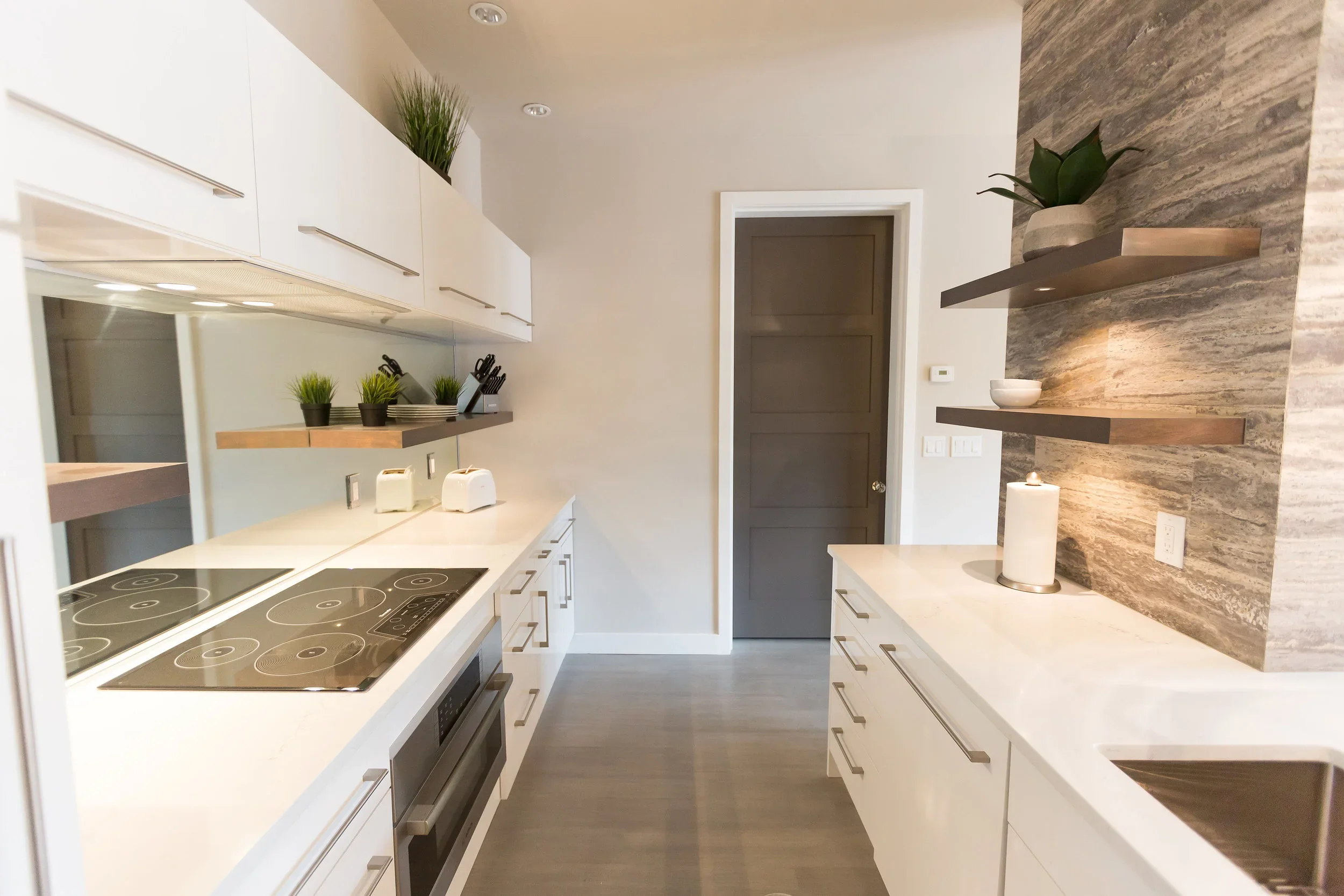 A modern kitchen with white cabinets, a white countertop, and a gray door. The wall behind the countertop has a textured stone finish with floating wooden shelves holding a potted plant and a bowl. There are small potted plants and kitchen appliances
