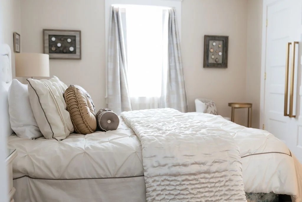 Bright bedroom with a bed dressed in white linens, decorative pillows, and a cream quilt. Light filters through sheer curtains on a window, flanked by framed wall art. A nightstand and small side table are visible.