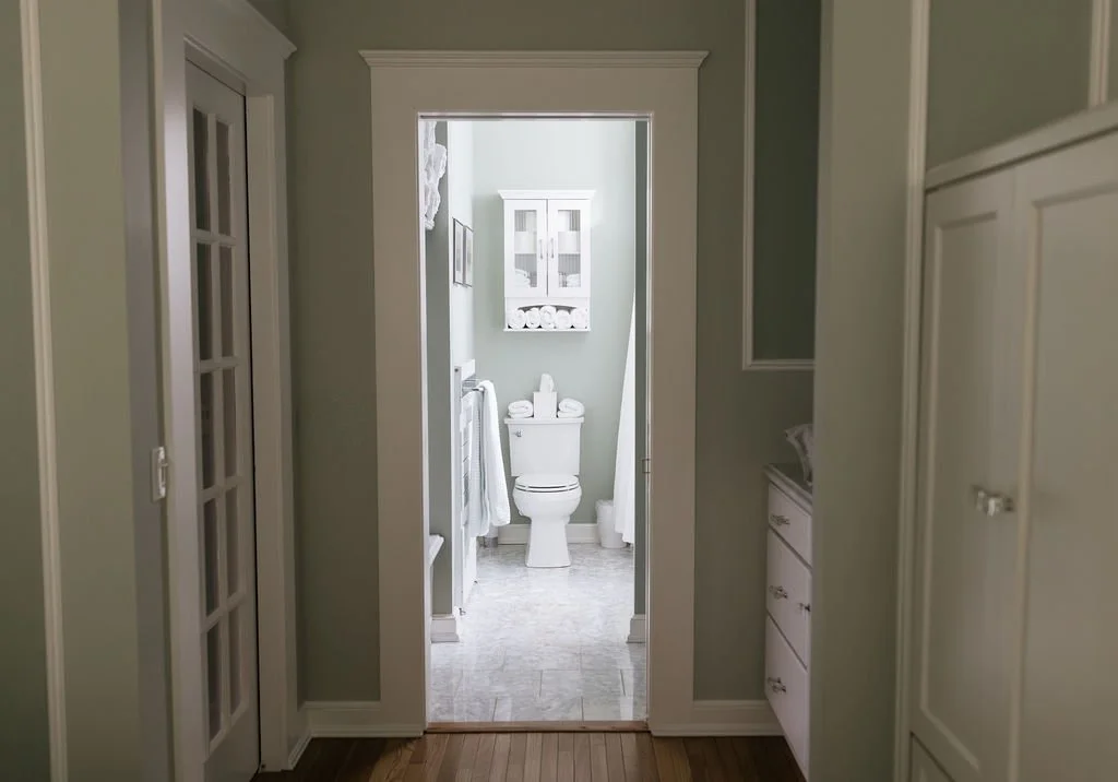 View of a small bathroom with a toilet, white cabinet above, and toiletries on top, seen through a doorway from a hallway with cabinets on the right and a door with glass panes on the left.