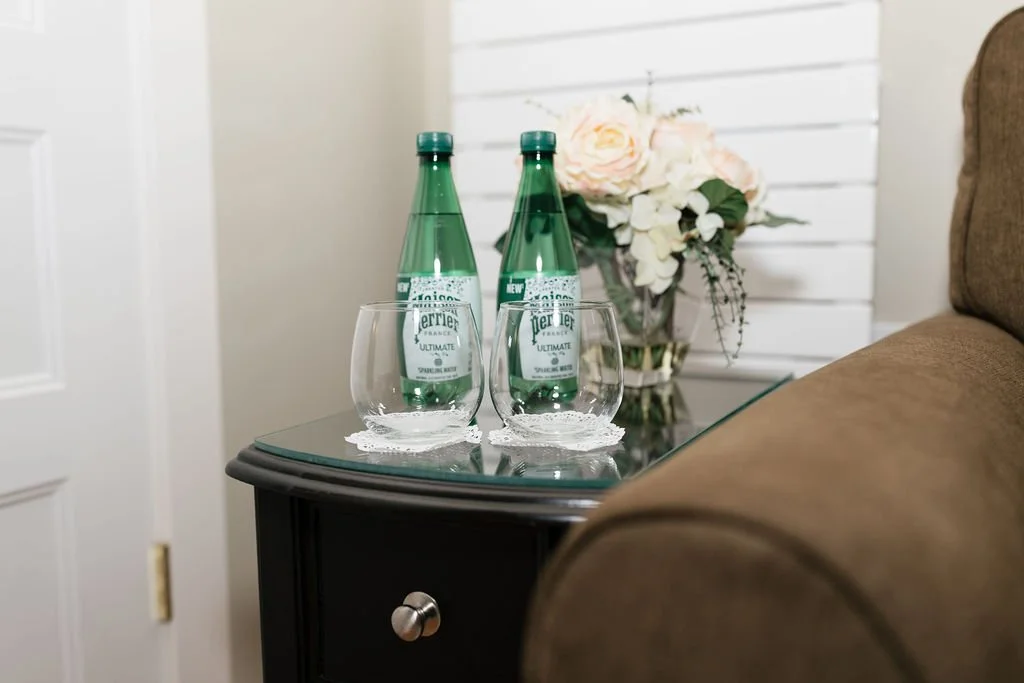 Two bottles of San Pellegrino sparkling water and two empty glasses on a glass-topped black table, with a flower arrangement in a vase in the background next to a white wall and part of a brown armchair.