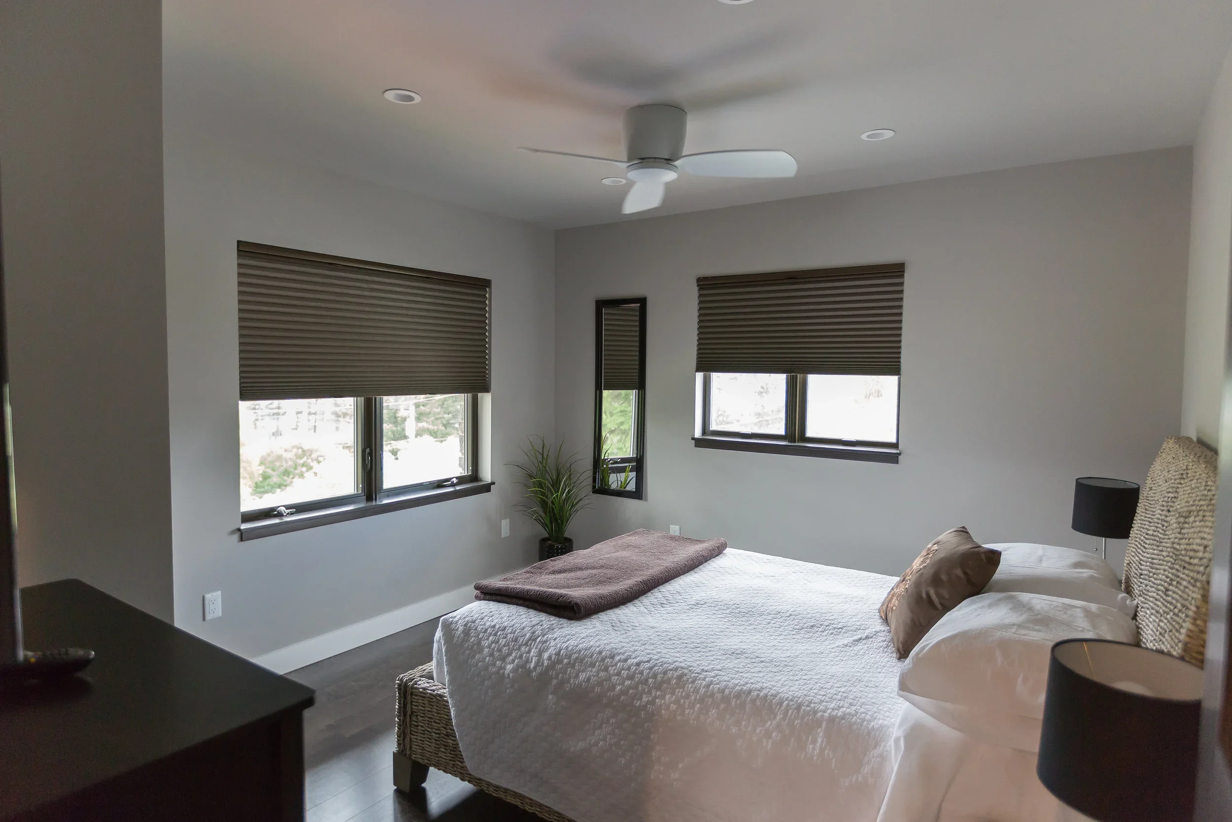 A bedroom with two windows covered by brown blinds, a bed with white bedding and brown pillows, a bedside table with a black lamp, a standing mirror, and a ceiling fan with white blades.