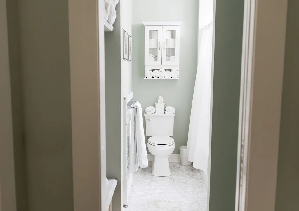 Small bathroom with a white toilet, white cabinet above it containing towels, and a white curtain on the right side, with windows allowing natural light in.