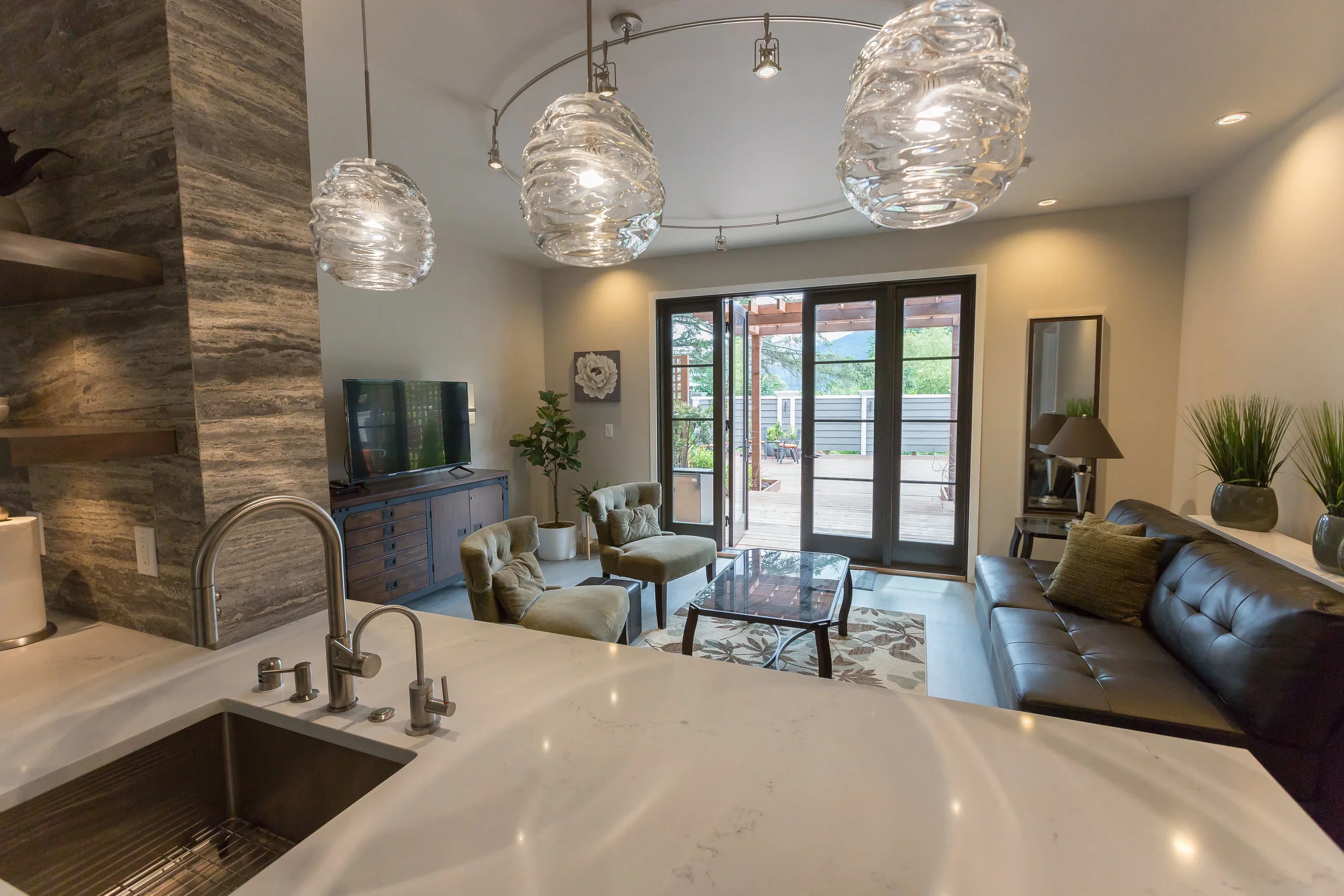 Living room with large glass doors opening to a deck, modern light fixtures, a flat-screen TV, plants, and a sitting area with chairs and a sofa.