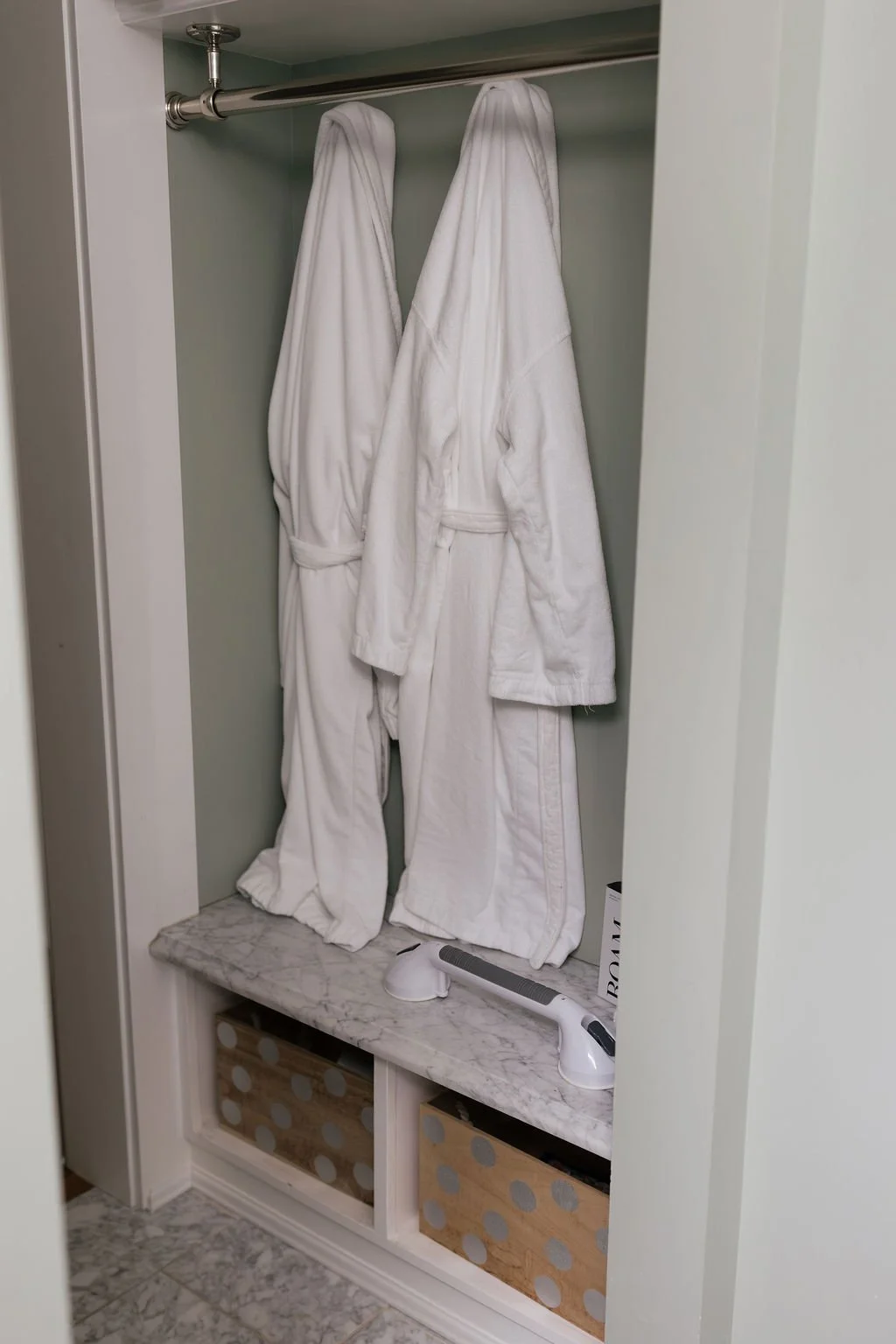 Two white bathrobes hanging on a metal rod inside a small closet with a marble shelf and storage bins below. There is a handheld vacuum on the shelf.
