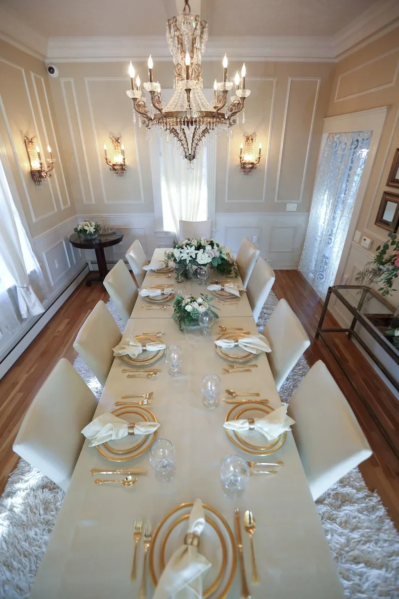 Elegant dining room with a long table set for a formal meal, decorated with floral centerpieces, gold-rimmed plates, white napkins, and gold flatware, illuminated by a large chandelier and wall sconces.