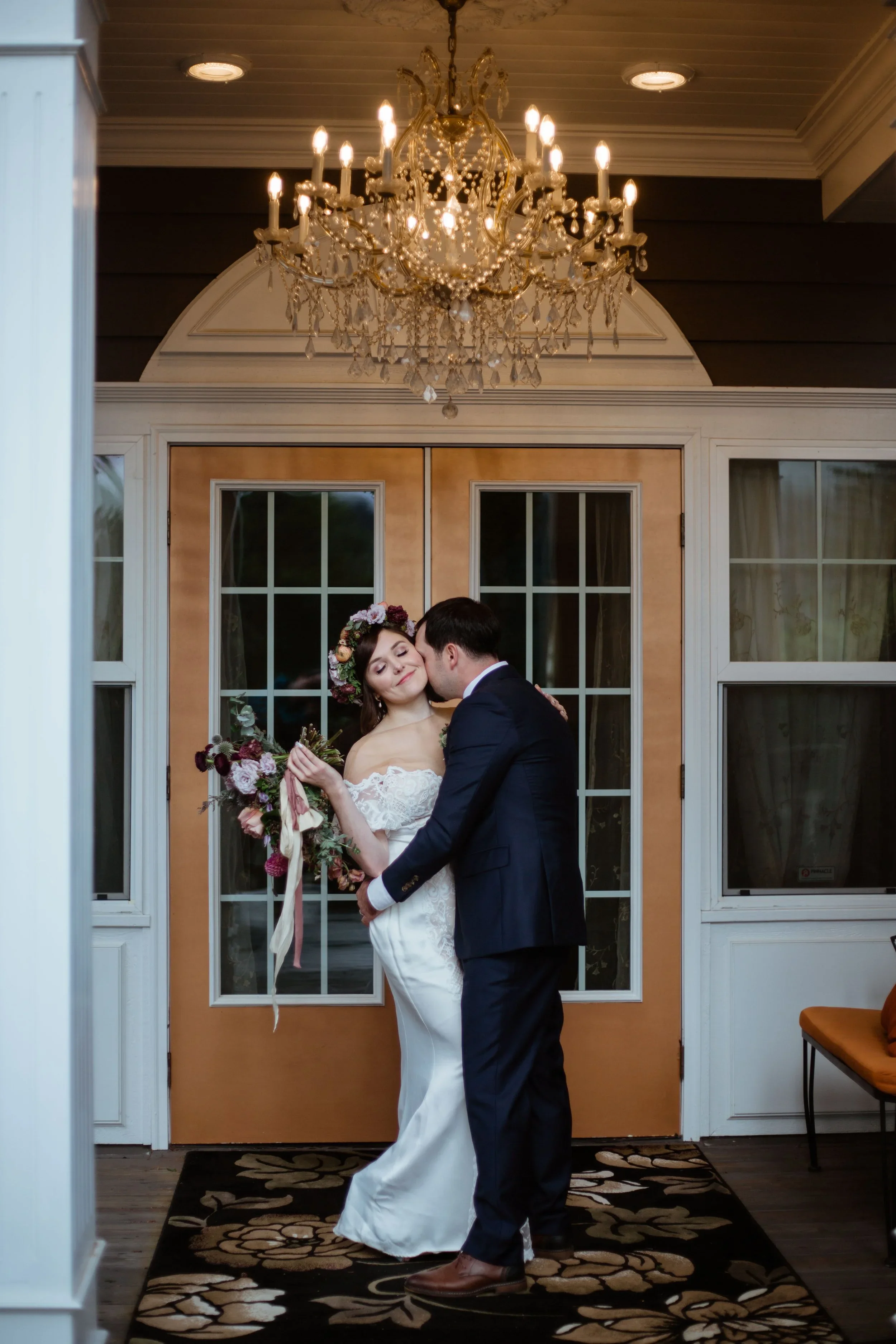 A bride and groom embrace and smile during their wedding in a warmly lit room. The bride holds a bouquet of flowers, and the groom wears a dark suit. A glamorous chandelier hangs from the ceiling, illuminating the scene.