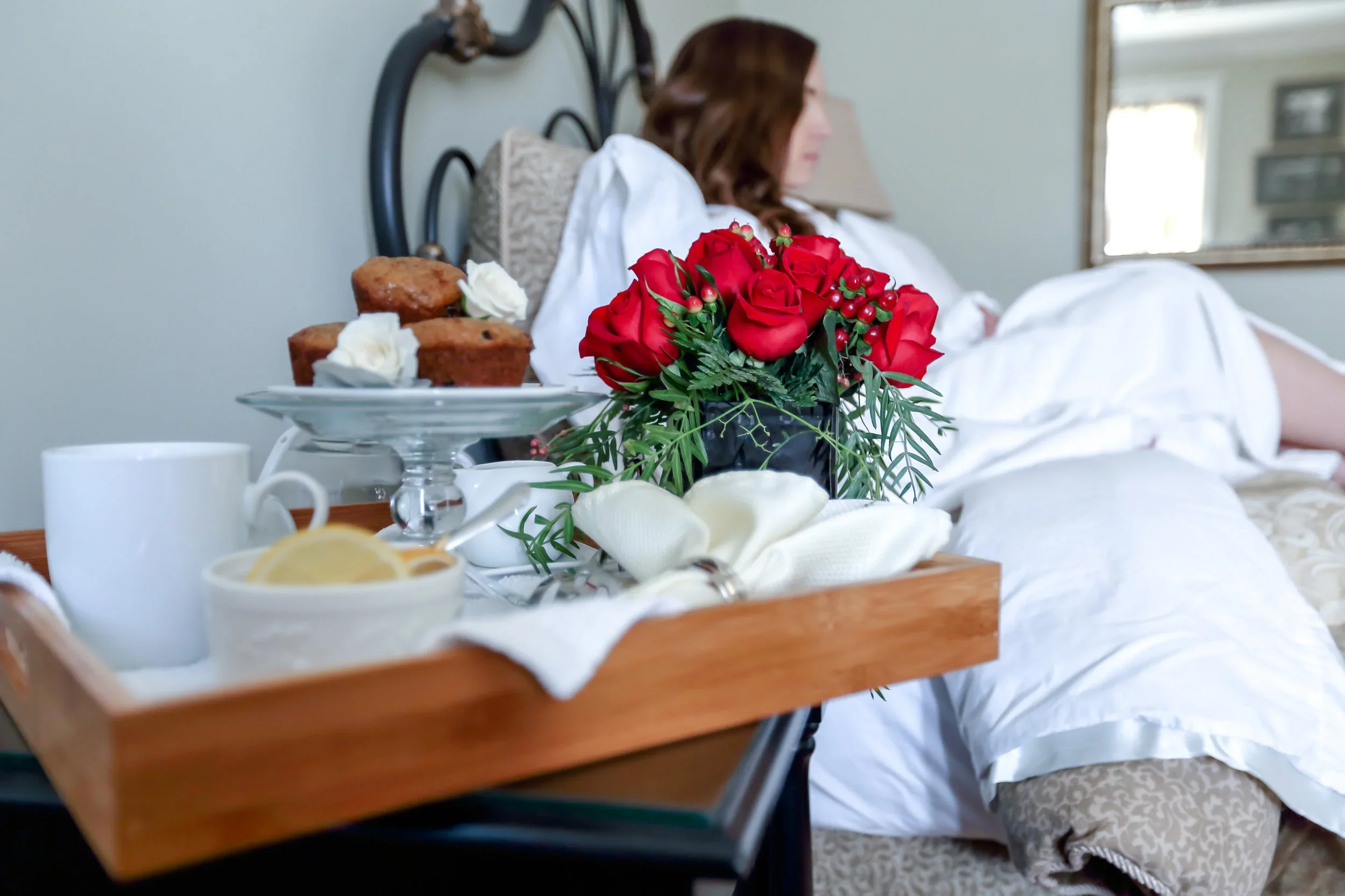 A bed with white bedding and a woman reclining against pillows, a tray with breakfast items including muffins, lemon slices, a white cup, and a flower bouquet of red roses and green foliage on the bedside table.