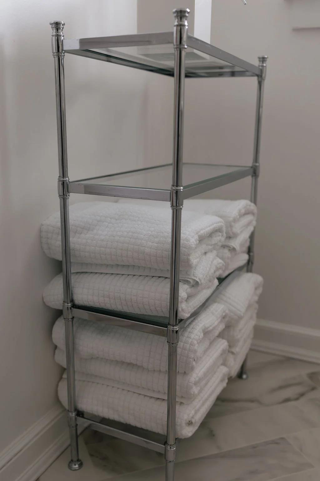 A metal shelving unit with three tiers of folded white bath towels, located next to a white wall and on a marble floor.