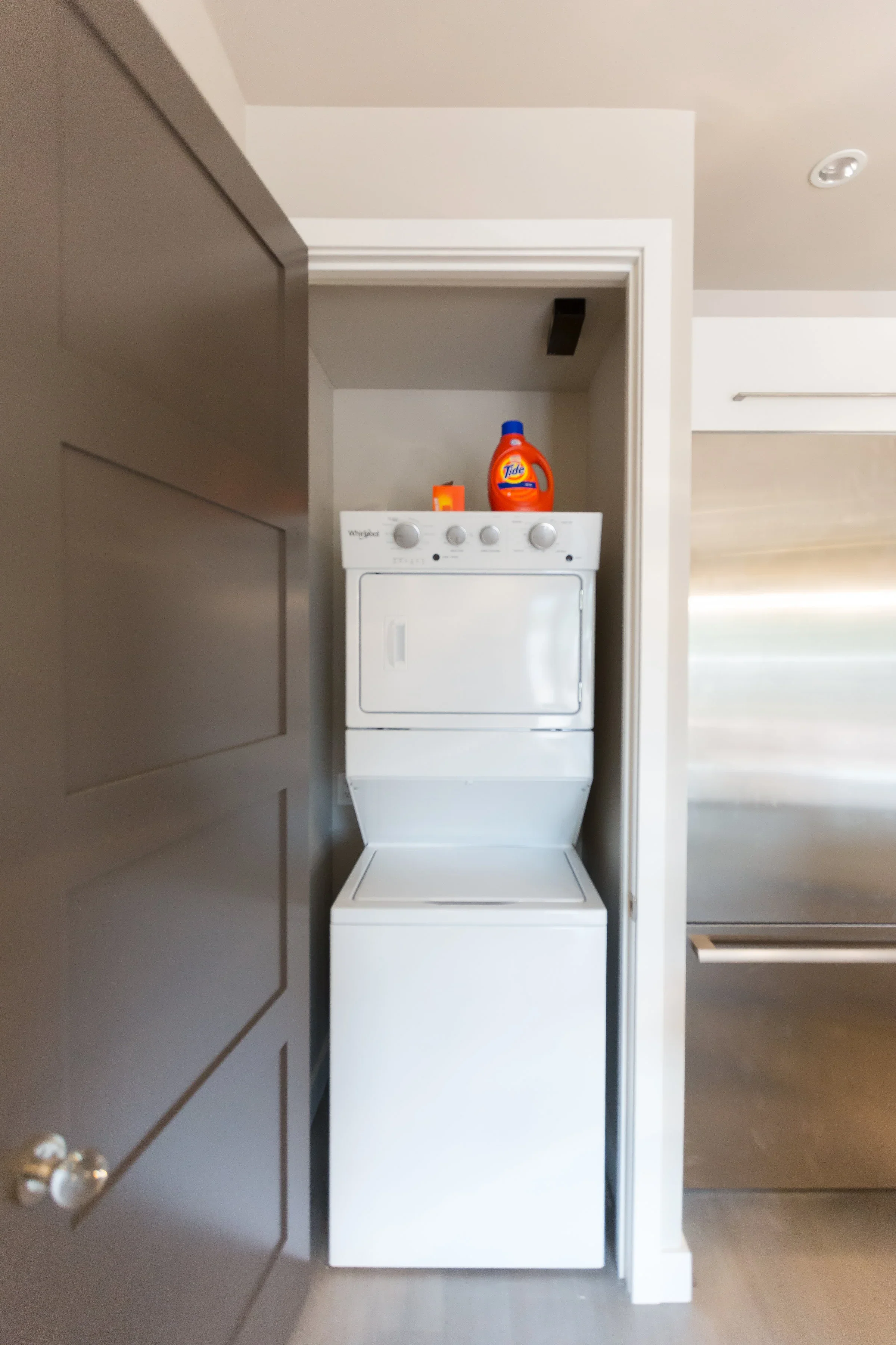 A laundry area with a stacked washer and dryer in a small niche. On top of the washer, there is a bottle of Tide laundry detergent and a small container. The area is enclosed by white wall trim and surrounded by a neutral-colored wall.