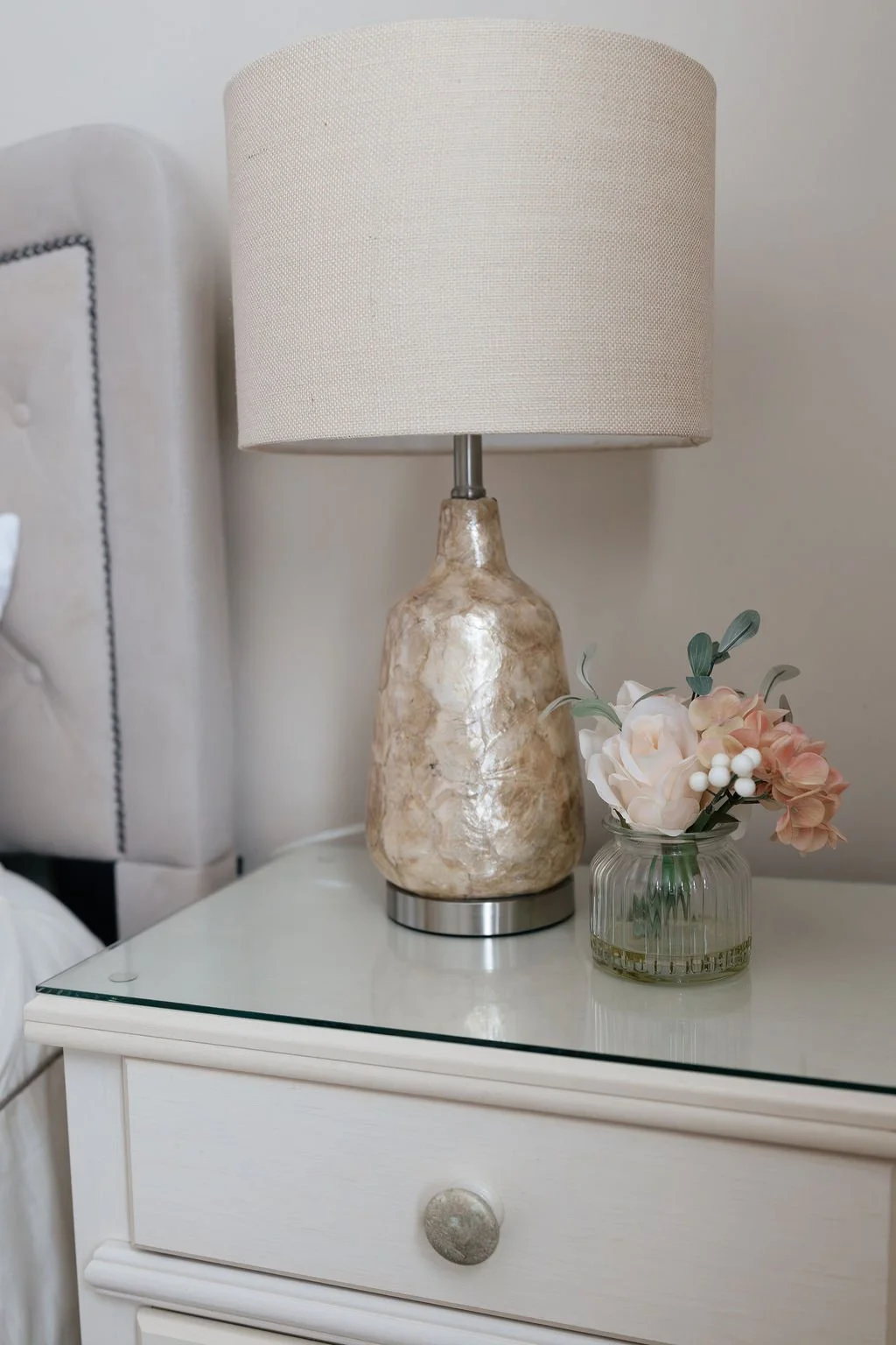 Lamplight on a white nightstand next to a glass vase with pink and white artificial flowers.