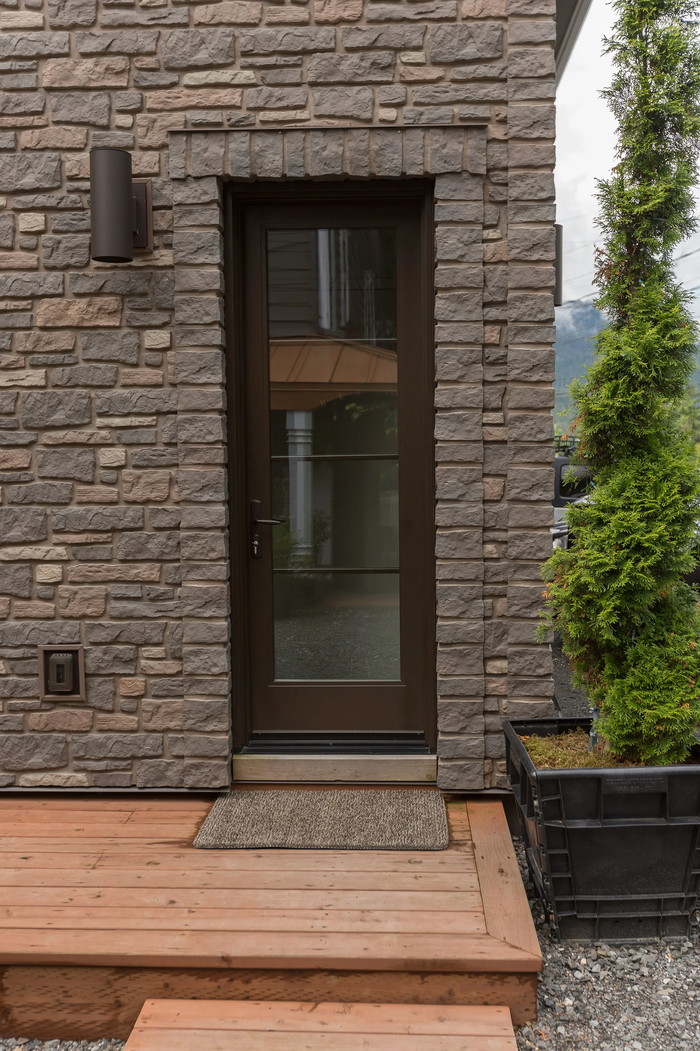 The image shows the front door of a modern house with exterior brick walls. A light fixture is mounted on the wall to the left of the door. There is a gray doormat in front of the door and a wooden deck leading to it. To the right, a potted shrub or 