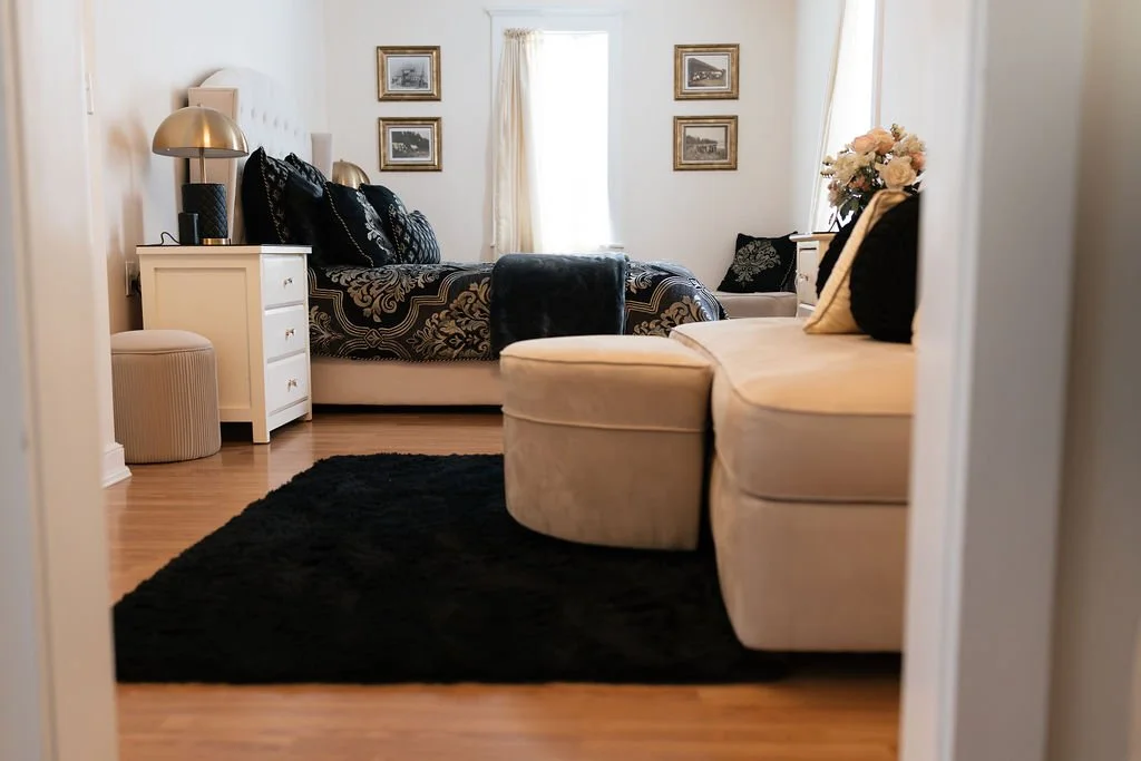 A cozy bedroom with a beige sofa, black and cream pillows, a white nightstand with a lamp, a bed with black and white bedding, a side table with fresh flowers, and framed pictures on the walls.