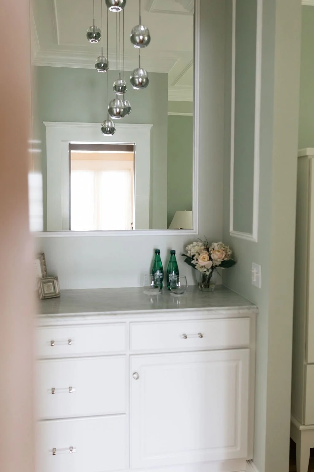 A bathroom vanity area with a large mirror, a vase of white roses, two green glass bottles, and two wine glasses on the countertop.
