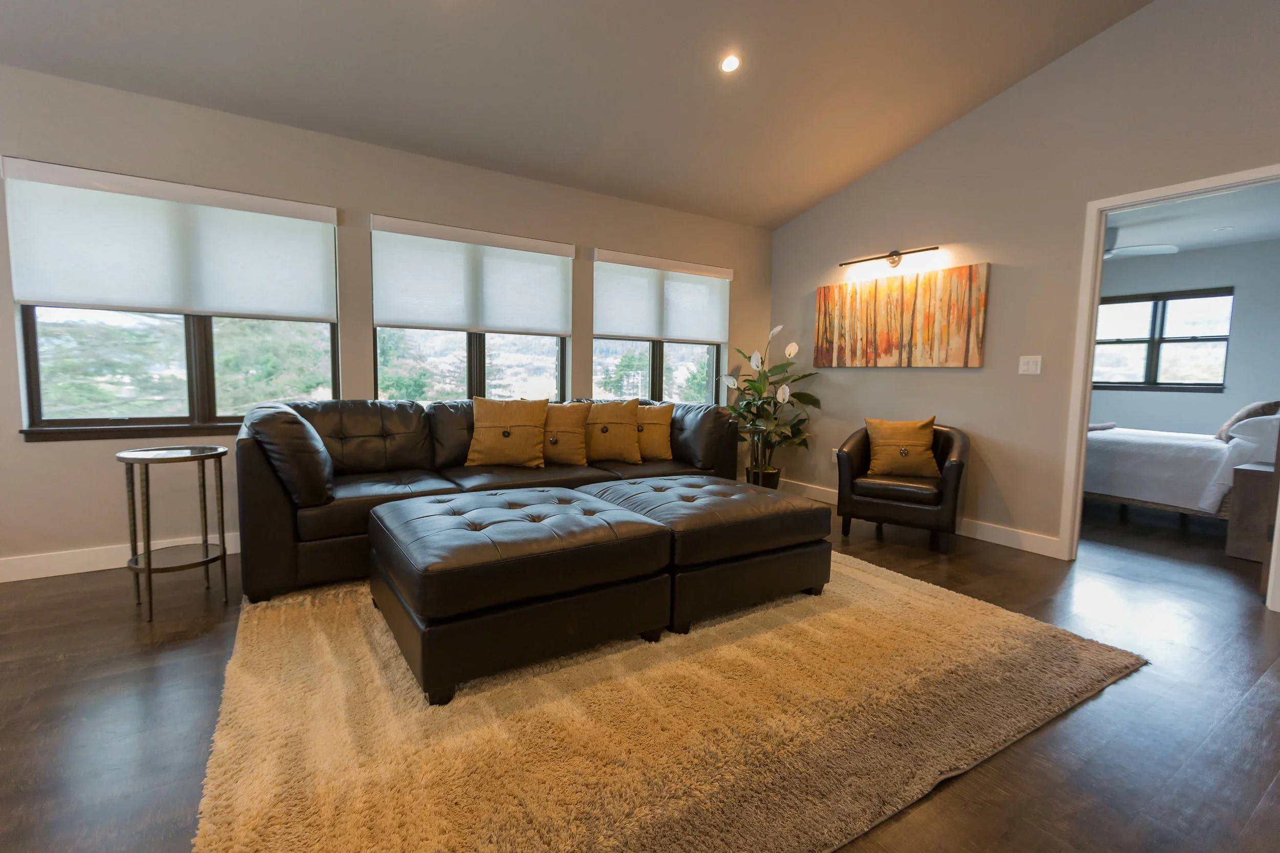 Living room with large sectional sofa, yellow pillows, a black armchair, a beige rug, a tall plant, and an abstract painting on the wall. Three large windows with white blinds provide natural light.