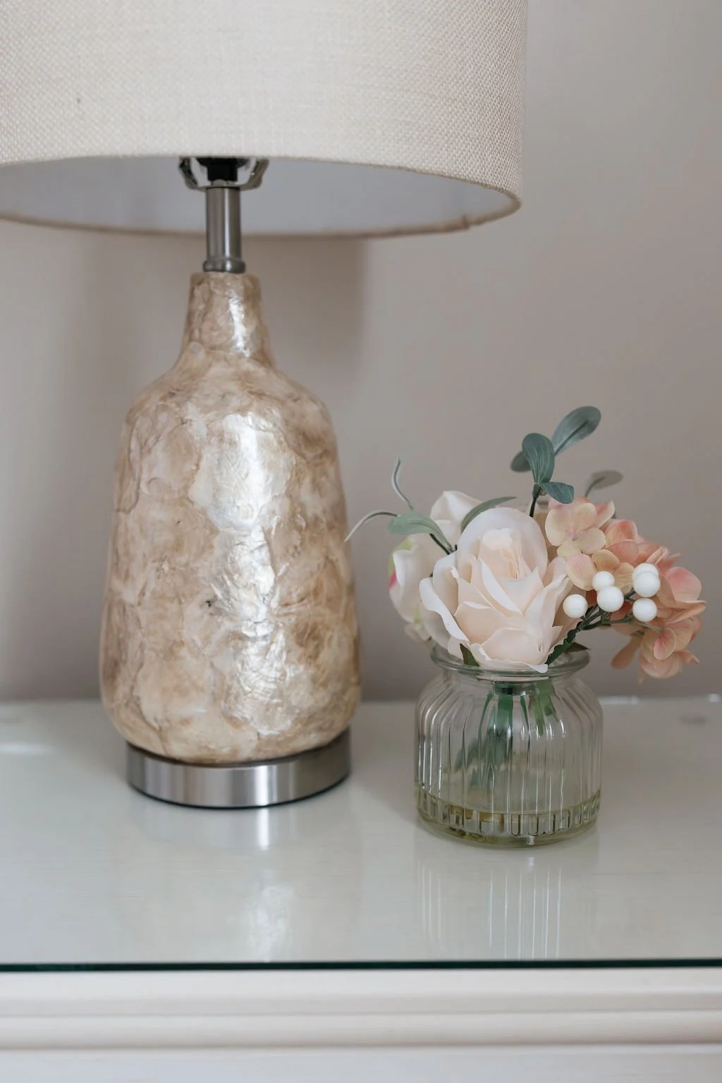 A beige marble table lamp with a linen shade next to a glass vase with white and pink artificial flowers.