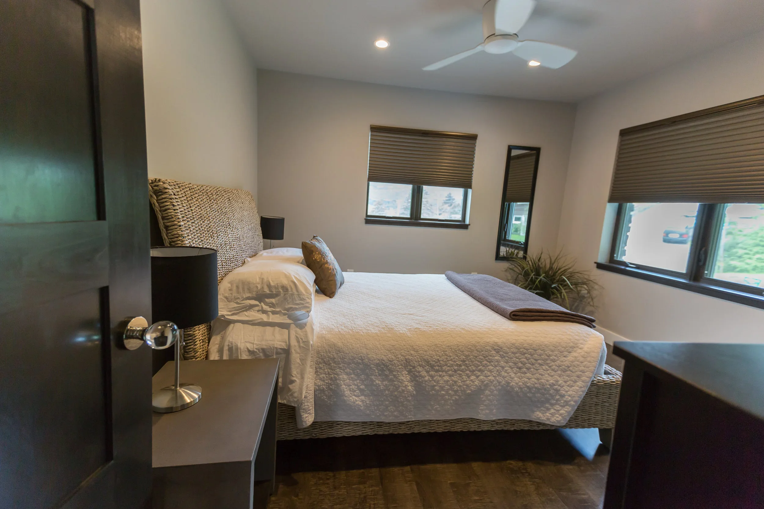 A cozy bedroom with a bed, multiple windows with closed blinds, a mirror, and a ceiling fan.