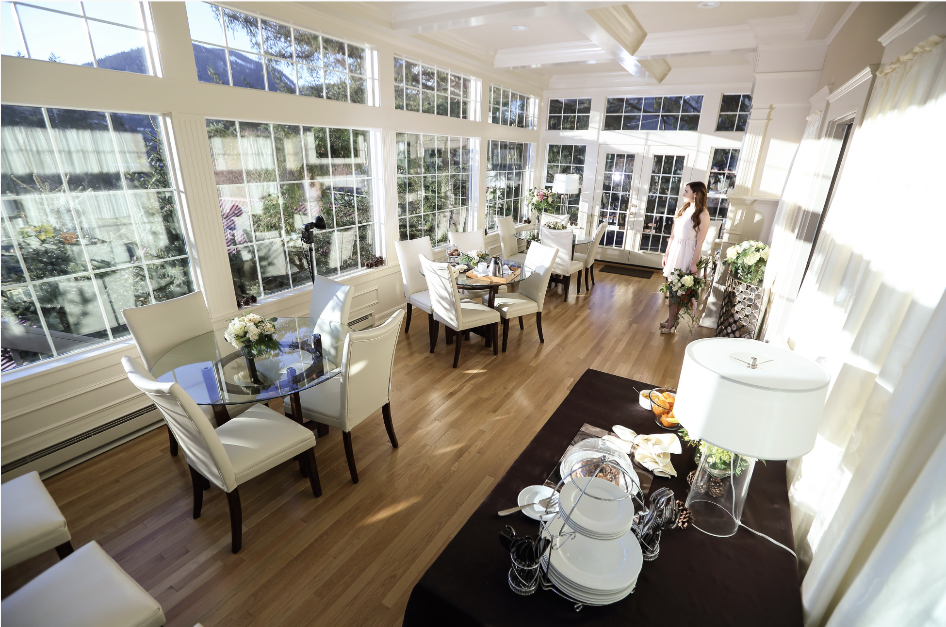 Bright dining room with large windows showing a view of trees and mountains, white chairs around glass tables, floral centerpieces, and a woman in a white dress standing near a decorated table with flowers and serving dishes.