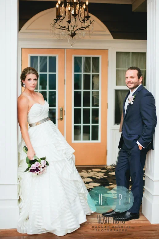 Bride in white wedding gown holding a bouquet of purple and white flowers, standing next to groom in a navy suit with a boutonniere, inside a porch with a chandelier and double glass doors in the background.