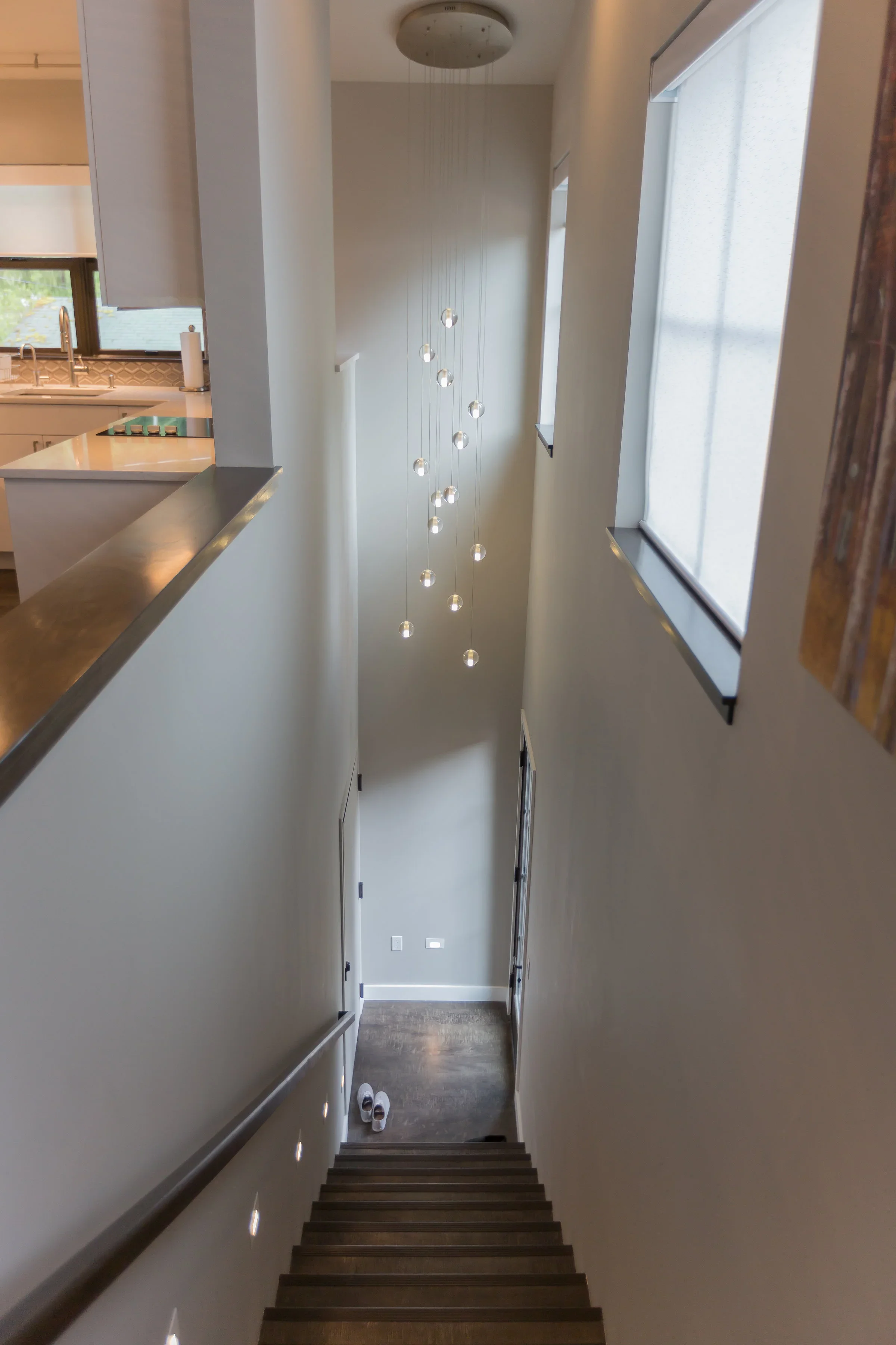 A view from the top of a staircase looking down into a hallway with white walls, wooden steps, and large windows on either side letting in natural light. A pair of shoes is at the bottom of the stairs, and a modern pendant light fixture hangs from th