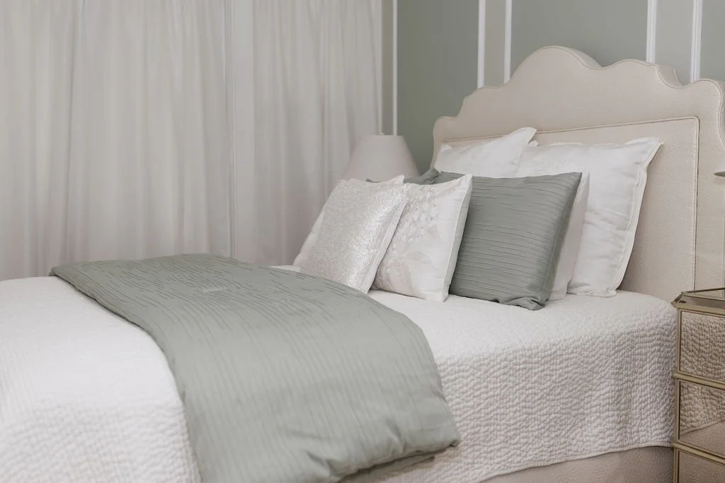 A neatly made bed with a white quilt and gray and white pillows in a softly lit bedroom with light-colored curtains.