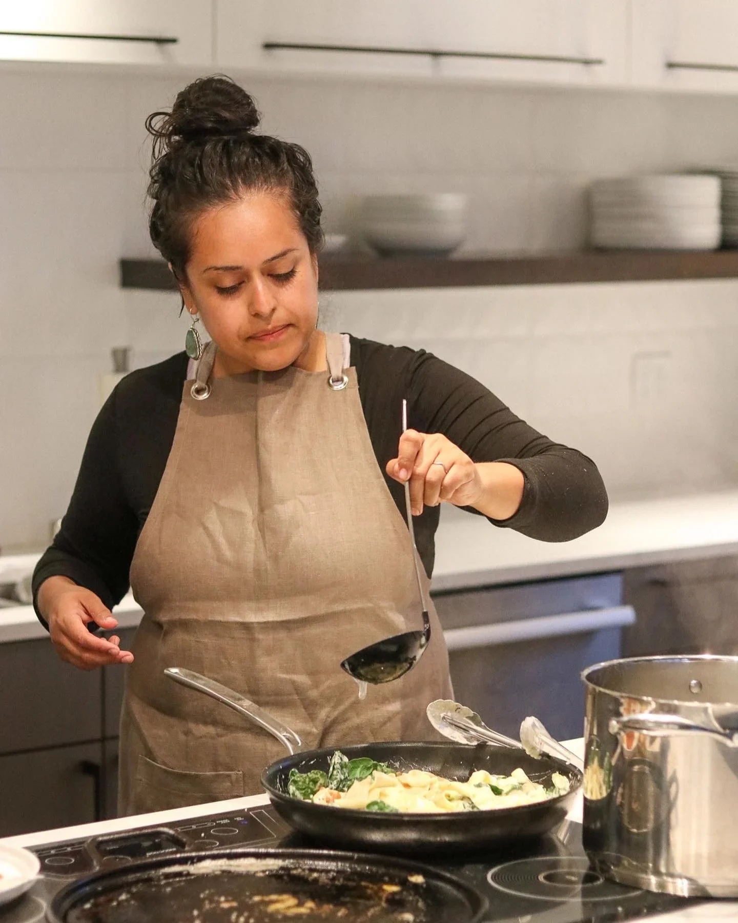 Woman cooking in a kitchen, wearing a beige apron and holding a ladle over a skillet of vegetables.