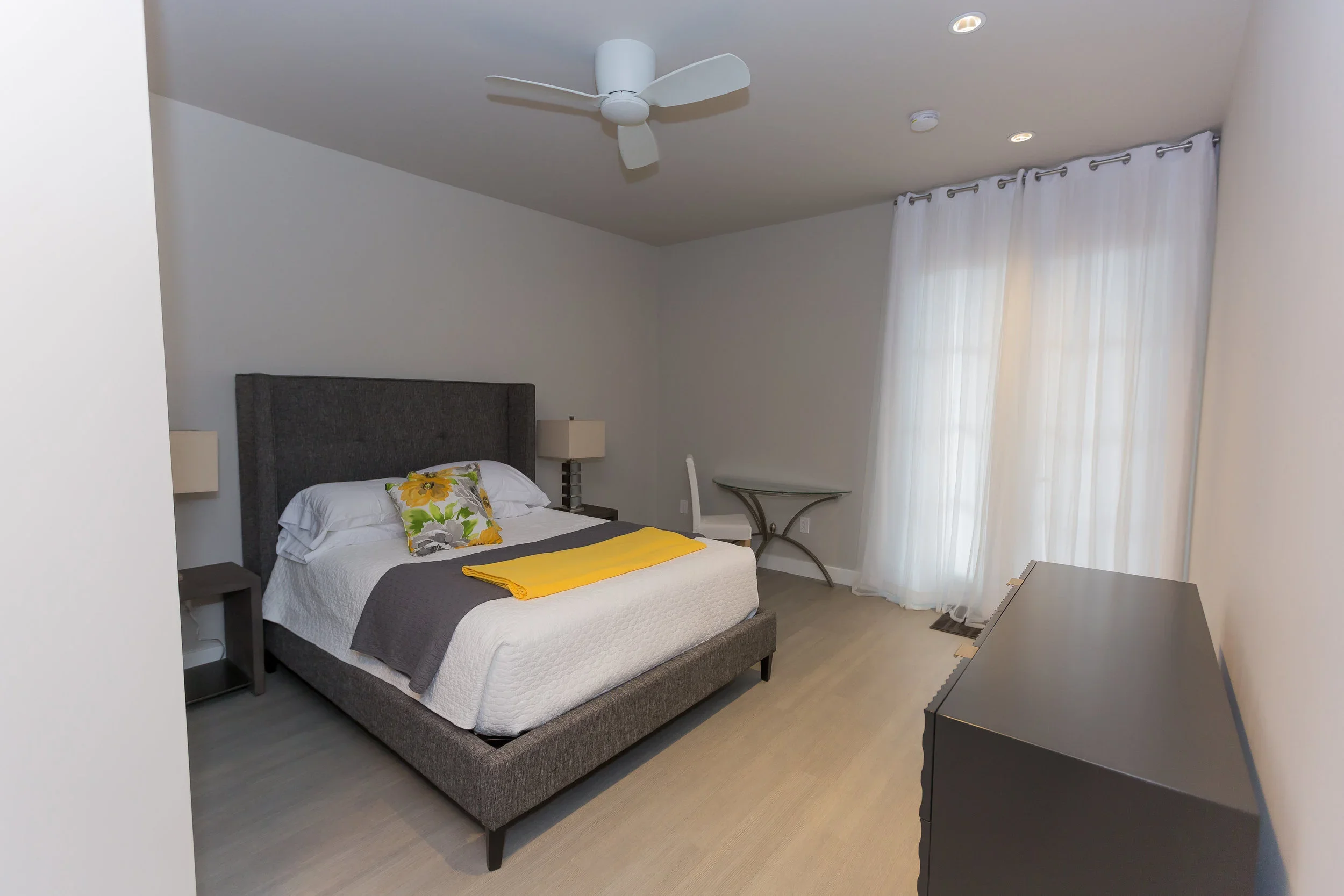 A bedroom with a gray upholstered bed, white bedding, and a floral pillow. There is a yellow blanket folded on the bed. The room has white walls, white curtains, a ceiling fan, and matching bedside lamps. A small table with curved legs is in the corn