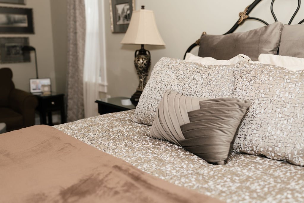 A neatly made bed with decorative pillows in a cozy bedroom, surrounded by side tables, lamps, and framed photographs.