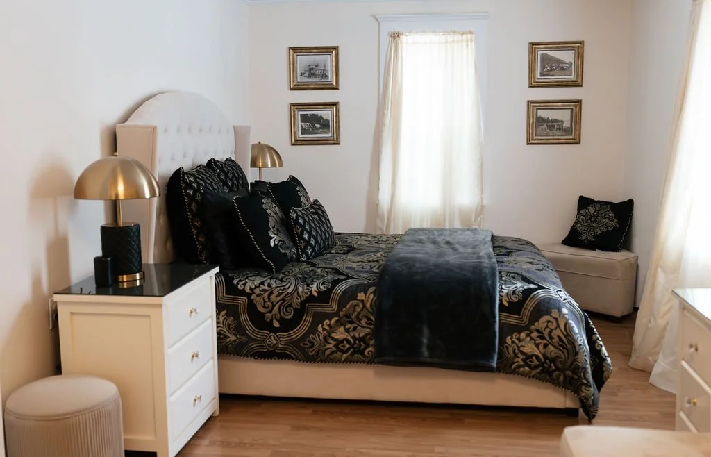 A bedroom featuring a bed with a black and gold damask-patterned duvet, black pillows, and a velvet black throw. There are two bedside lamps with gold shades on white nightstands, a beige bench at the foot of the bed, and framed black and white photo