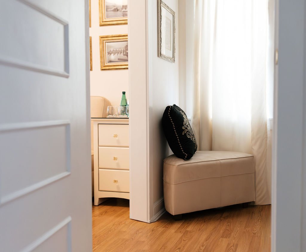 View of a cozy corner in a room with cream-colored curtains and a beige upholstered bench with a black decorative pillow outlined in gold placed on it.