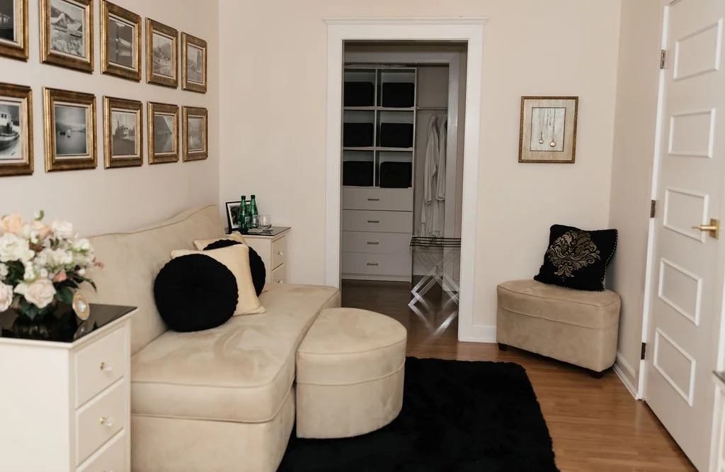 Living room with beige sofa, black round pillows, white side table with flowers, framed pictures on wall, black rug, beige chair with black and gold pillow, open closet in the background.
