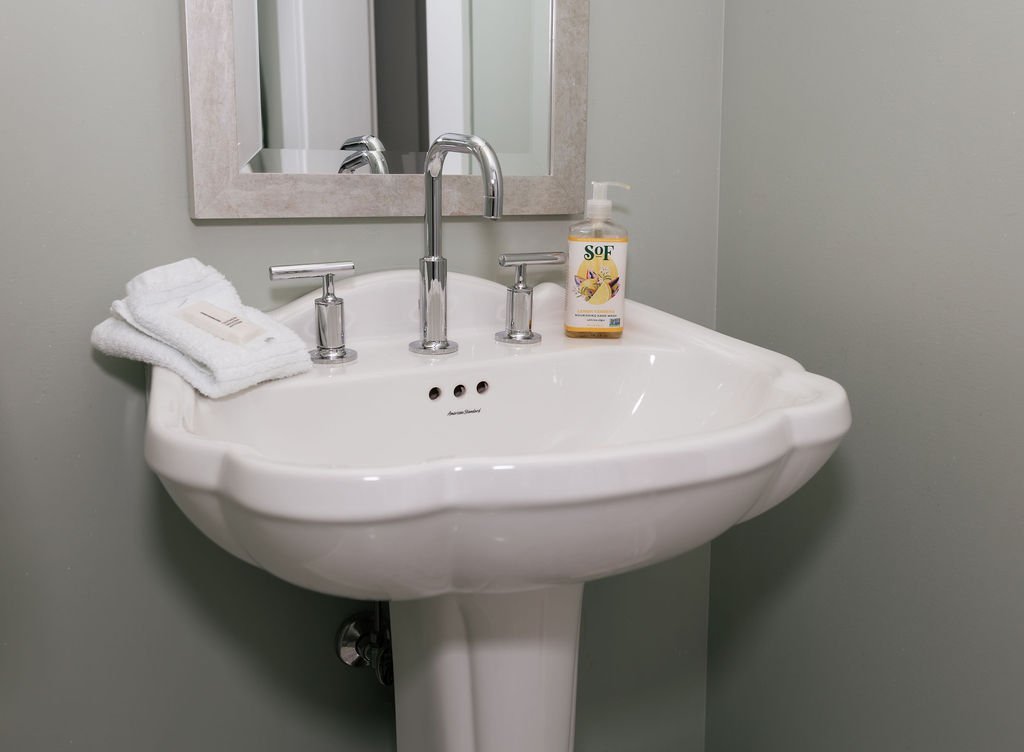 A white pedestal sink with a chrome faucet and handles, a folded white towel, a small soap dish, and a bottle of liquid soap in a bathroom, with a mirror above the sink.